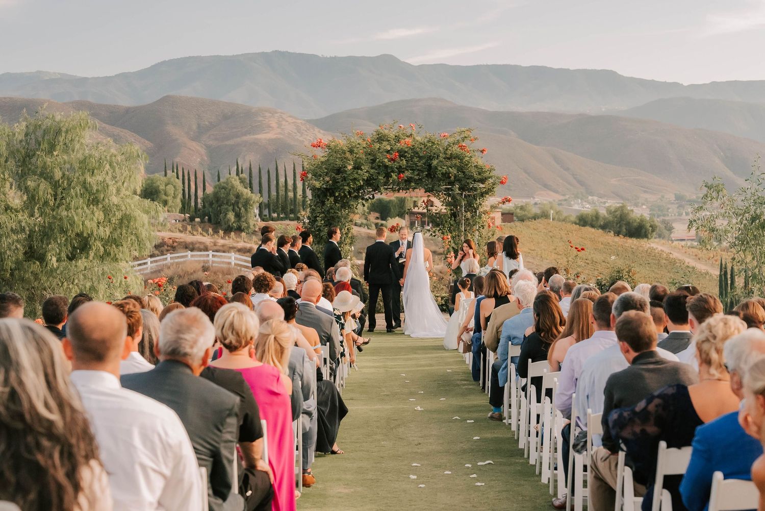 Outdoor wedding ceremony with guests seated in rows, couple at floral arch, vineyard and mountain backdrop.