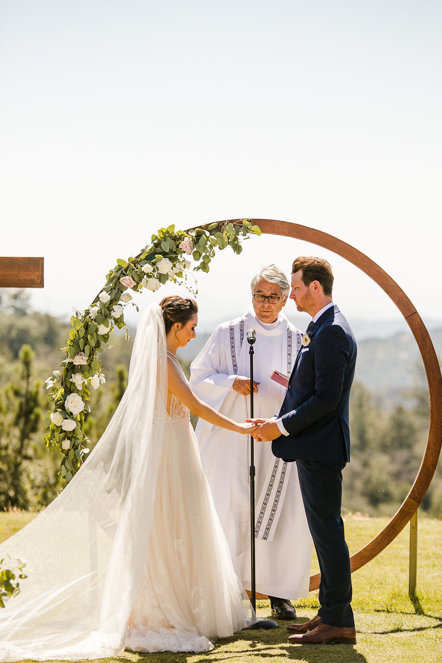 Bride and groom exchange vows under a circular wooden arch adorned with white flowers and greenery at an outdoor wedding ceremony.