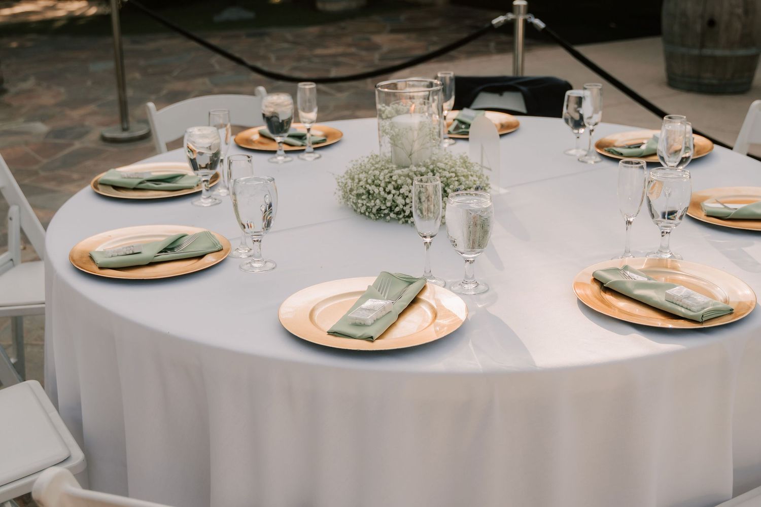 Elegant wedding reception table with white linen, gold chargers, sage green napkins, and a baby's breath centerpiece.