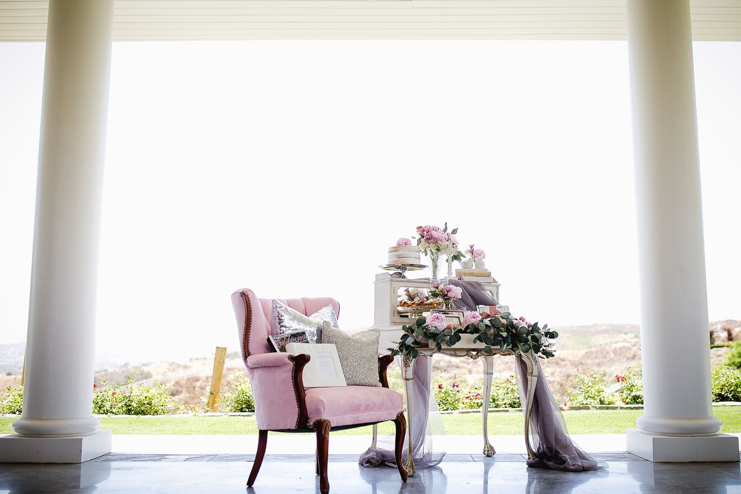 Pink wingback chair beside floral dessert table on elegant porch with white columns and garden view.