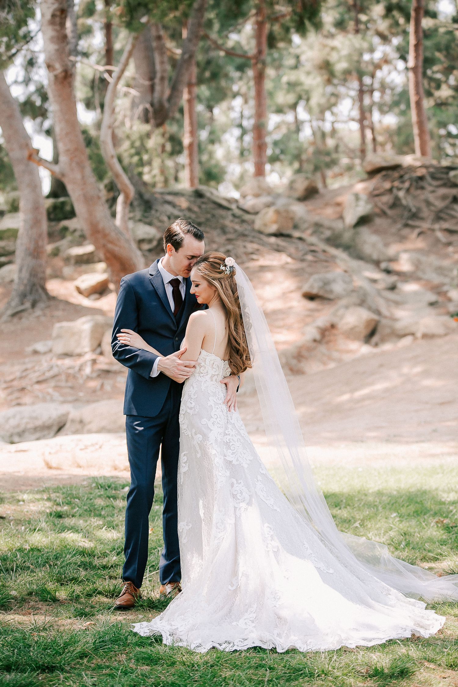 A bride in a lace gown and veil embraces a groom in a navy suit outdoors among trees and rocks.