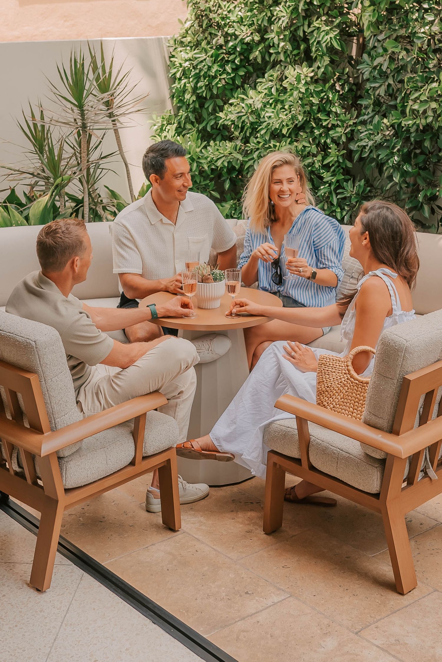 Four friends socializing on a stylish outdoor patio with wooden chairs, drinks, and lush greenery backdrop.