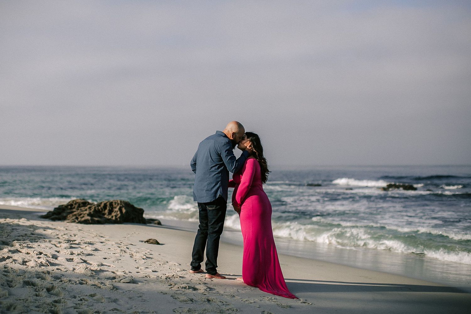 Couple kissing on sandy beach, pregnant woman in long pink gown with ocean waves behind them.