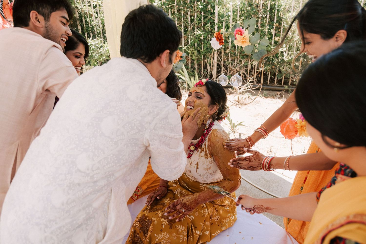 Family and friends applying haldi turmeric to seated bride during traditional Indian wedding ritual.