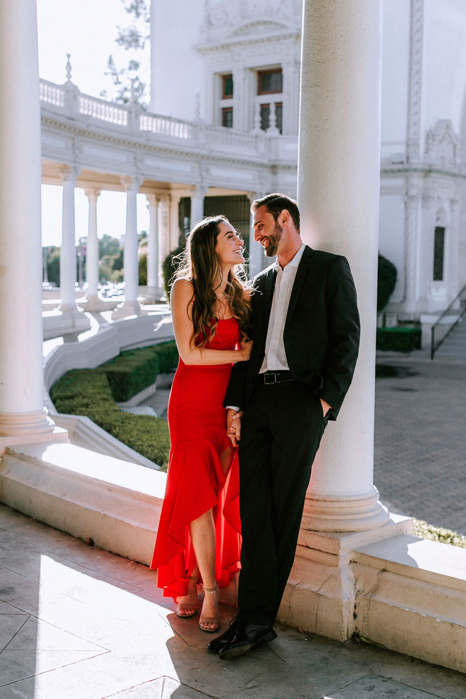 Couple in red dress and black suit leaning against white columns at historic building.
