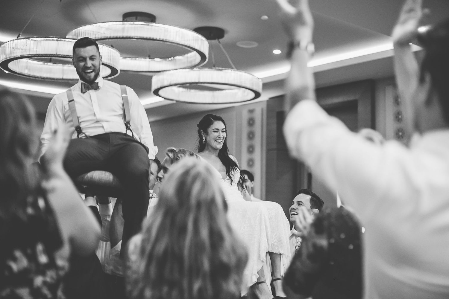 Black and white wedding reception photo of groom lifted in chair surrounded by cheering guests under circular chandeliers.