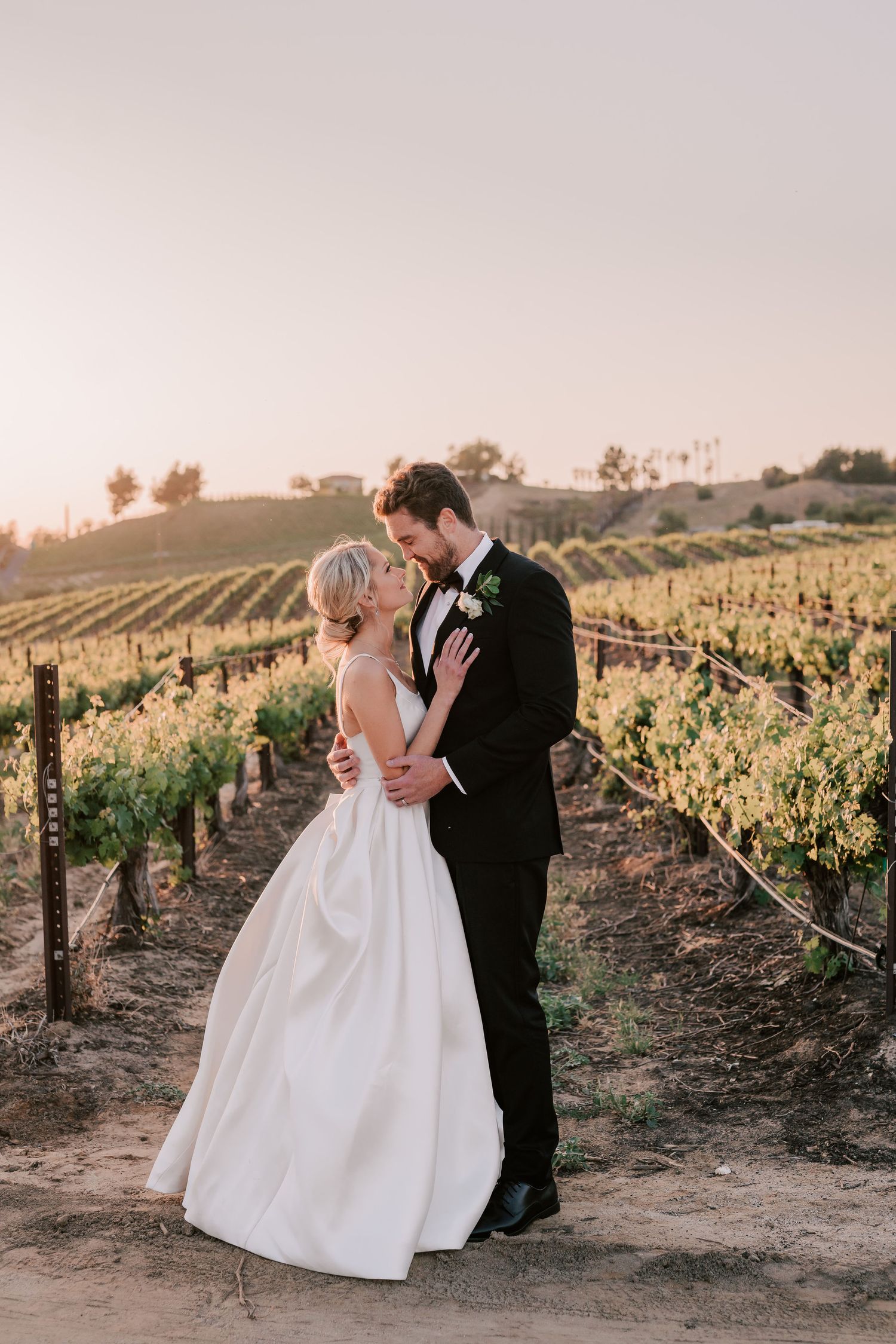 Bride and groom share a romantic kiss at golden hour in a scenic vineyard, bride in white gown, groom in black tuxedo.