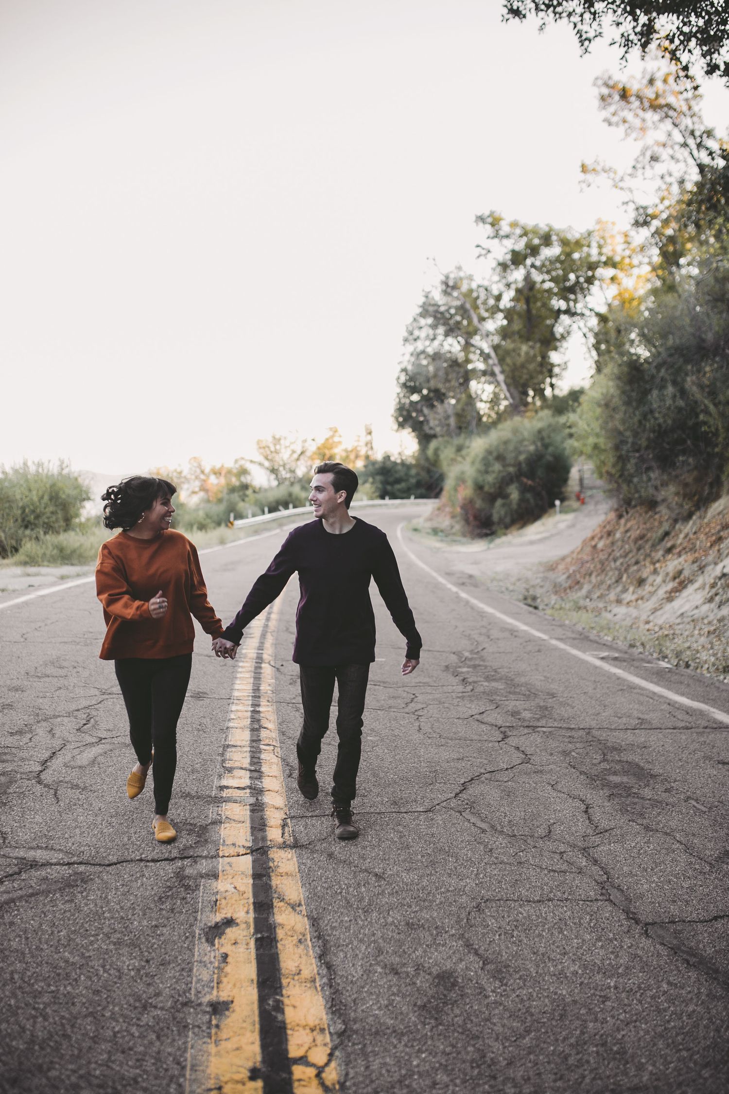 A couple holds hands while walking together down an empty road surrounded by trees.