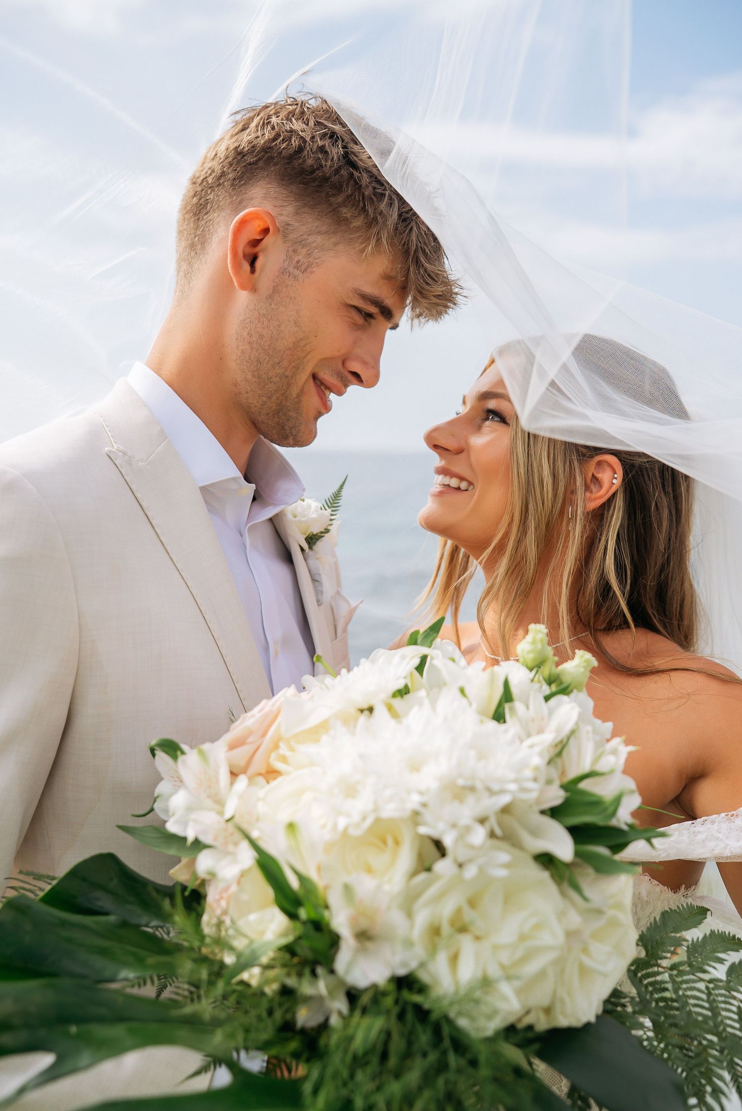 Smiling bride and groom share romantic moment under veil with white floral bouquet by the ocean.