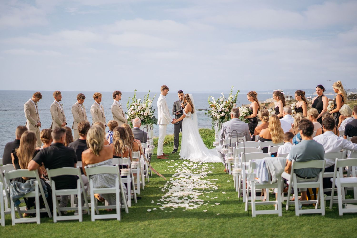 Outdoor wedding ceremony on oceanfront lawn with white chairs, floral arch, wedding party, and guests overlooking the sea.