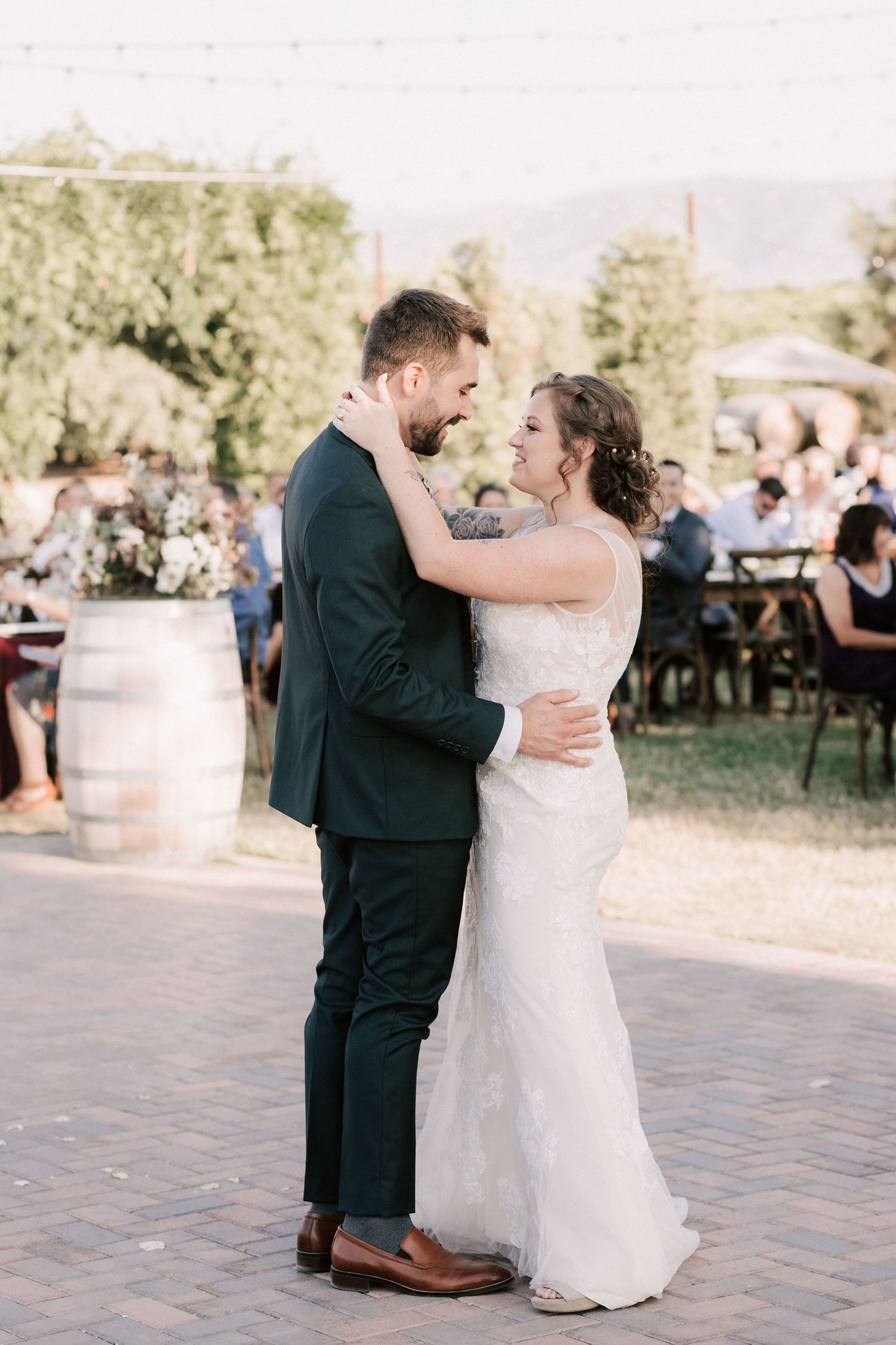 Bride and groom share first dance outdoors at winery wedding reception, surrounded by guests and barrel decorations.