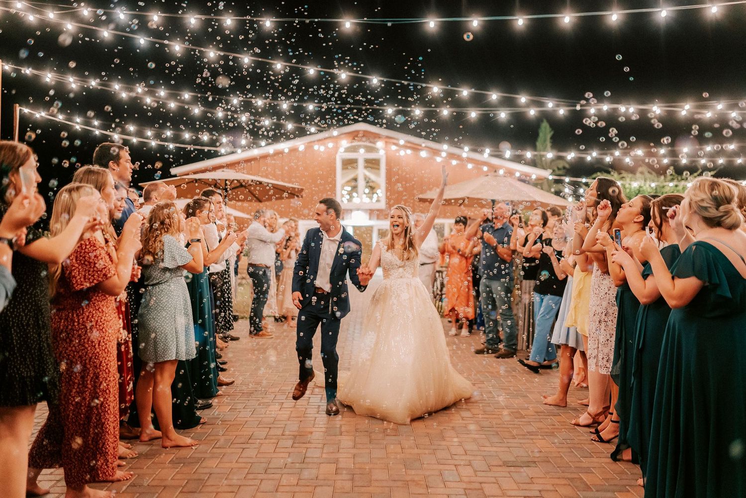 Bride and groom exit wedding reception under string lights surrounded by cheering guests in colorful attire.