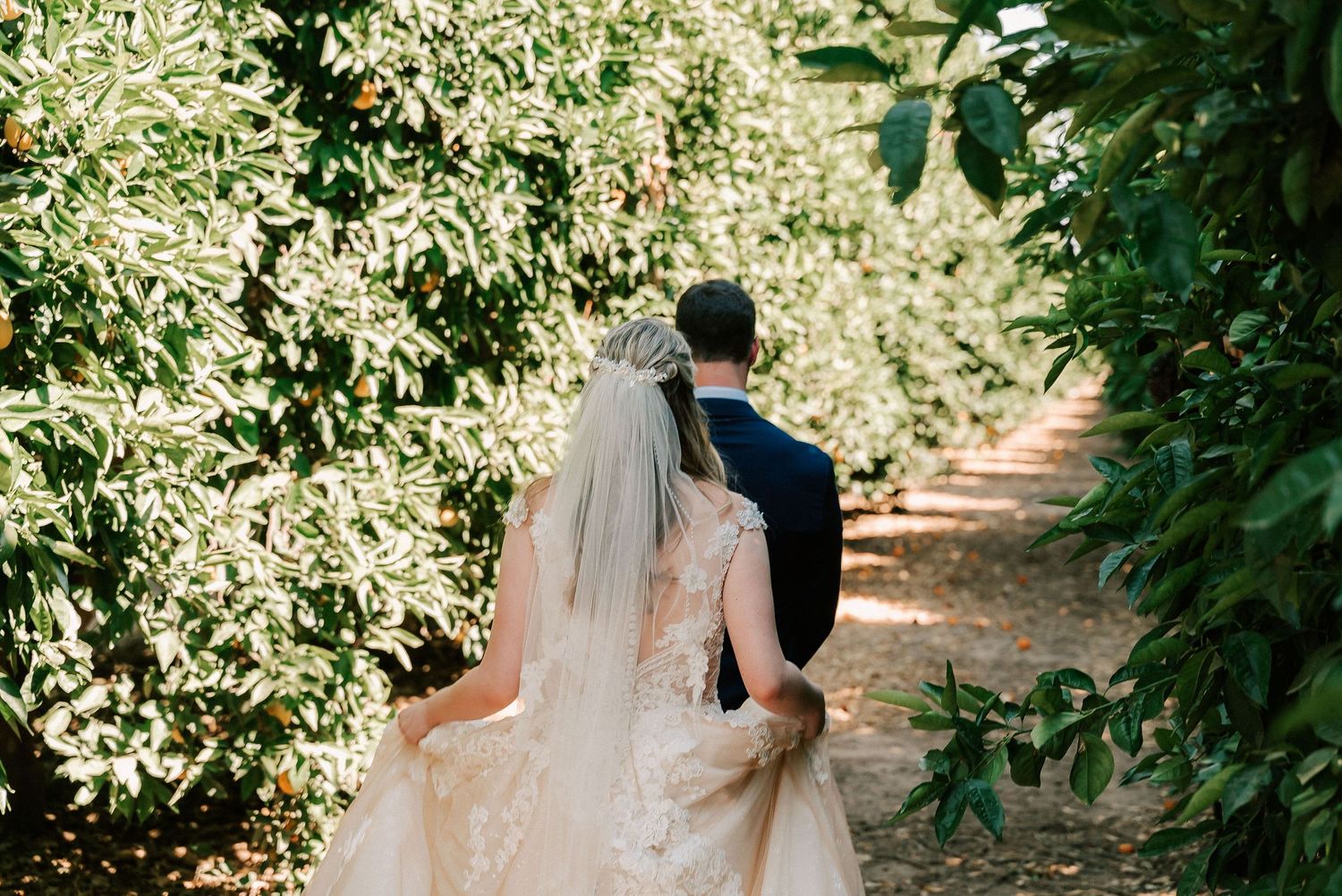 A bride and groom walk hand in hand through a lush green garden pathway on their wedding day.