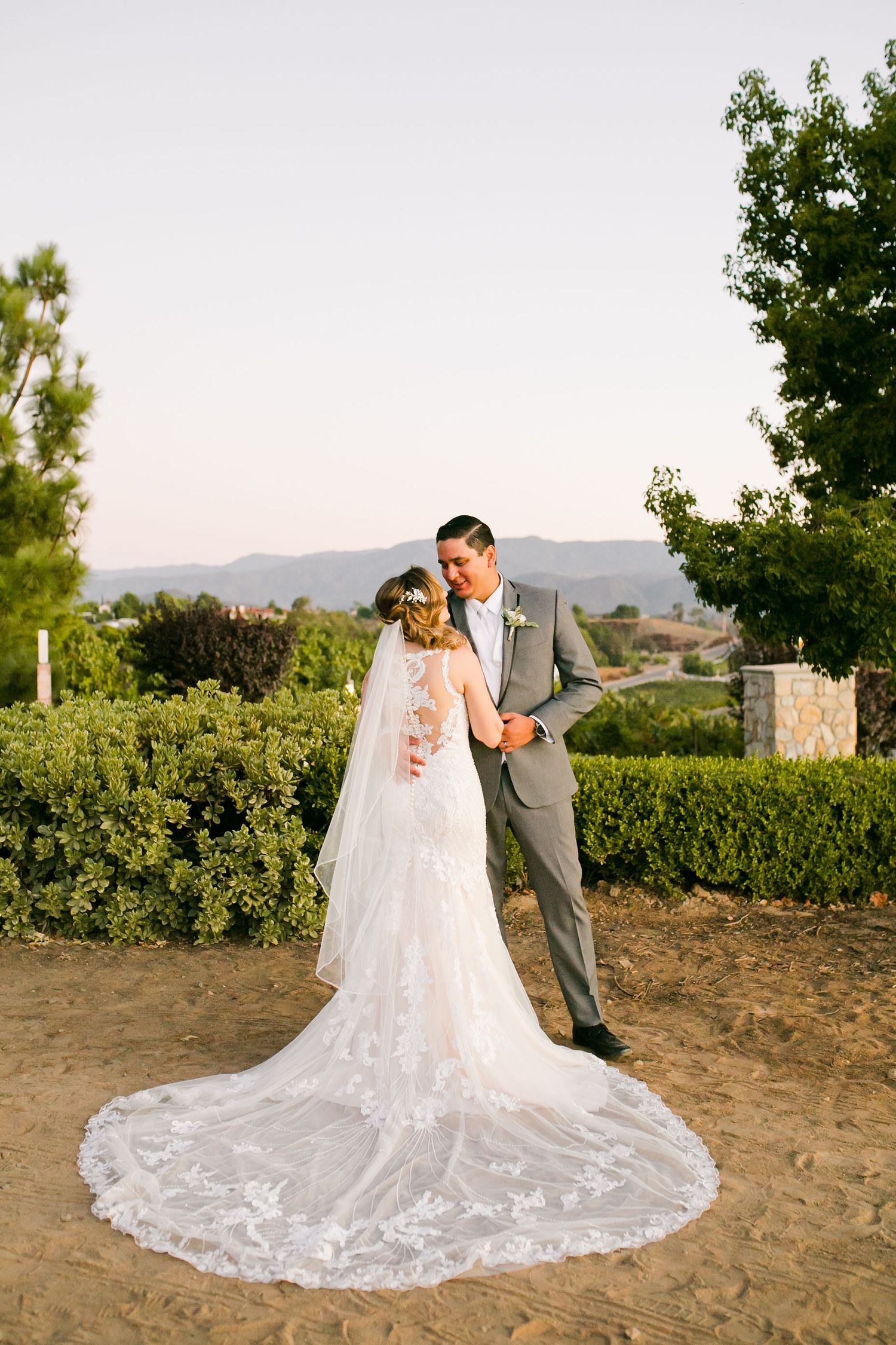 Bride and groom share a romantic moment outdoors, her lace cathedral veil trailing on sandy ground with green hedges behind.