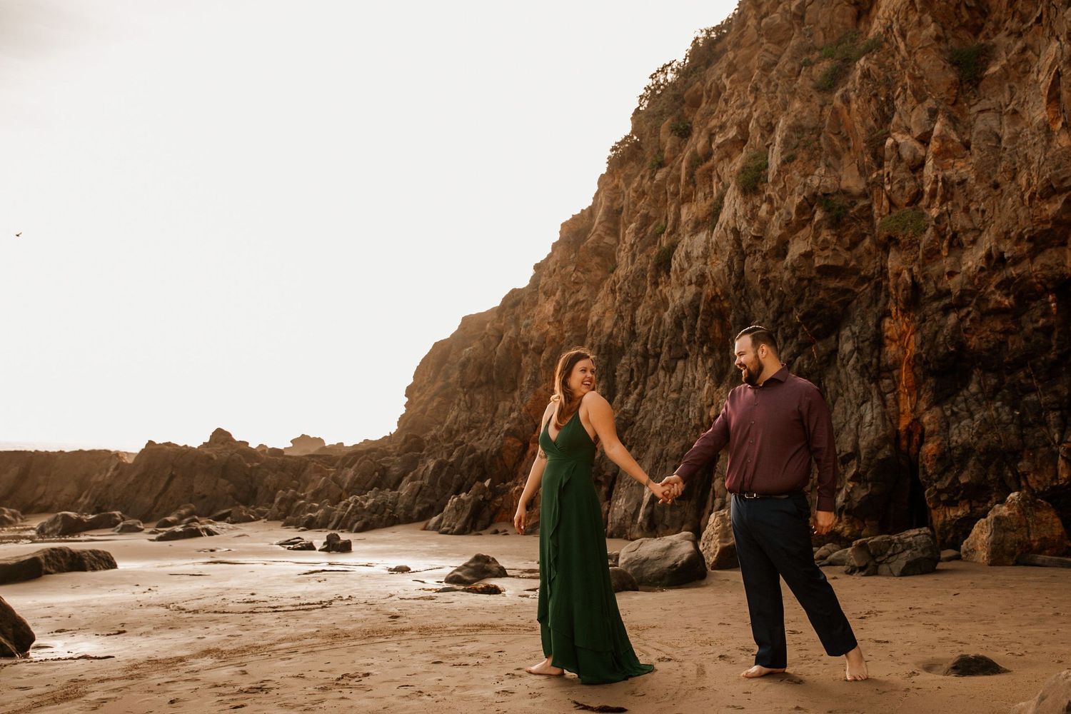 Couple holds hands on rocky beach, woman in green gown, man in burgundy shirt, dramatic cliffs behind them.