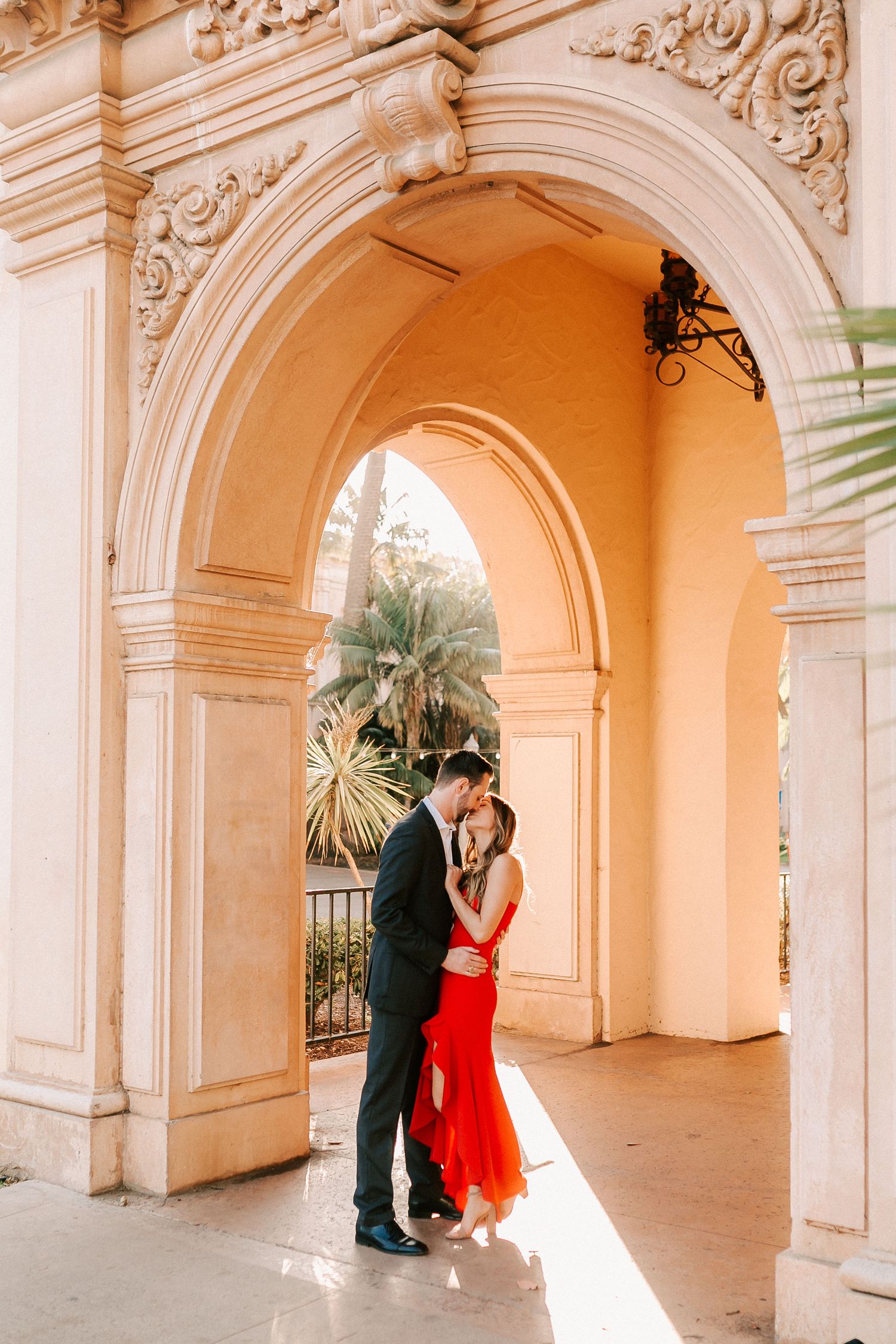 Romantic couple in red dress and dark suit embracing under ornate Spanish colonial arch at golden hour.