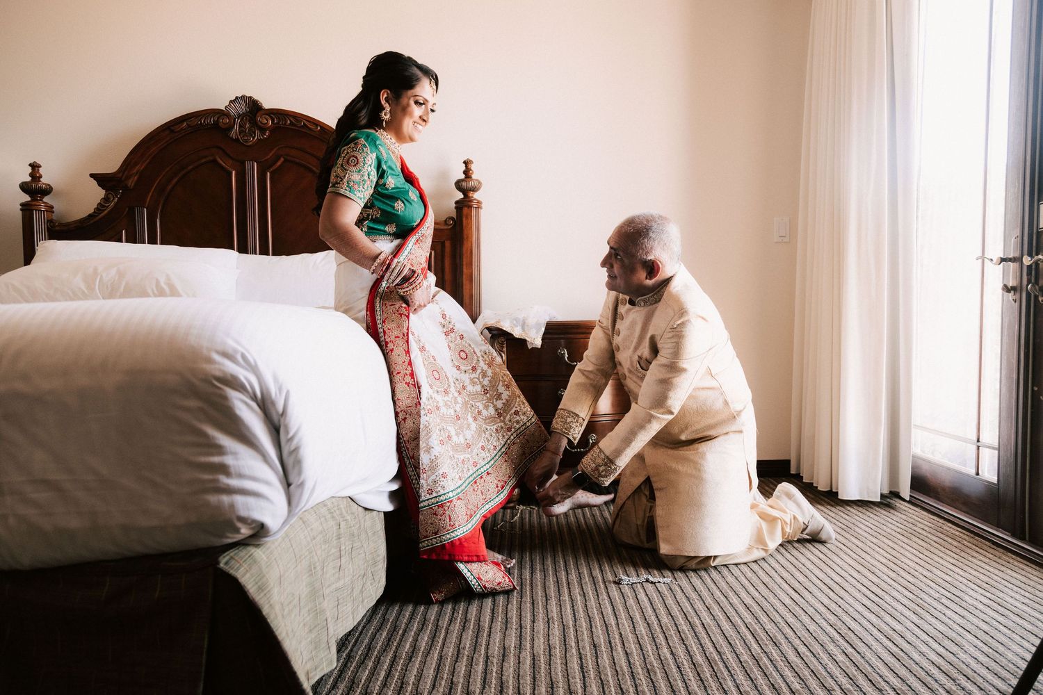 Indian bride in green saree getting shoes fastened by elderly man in hotel room.