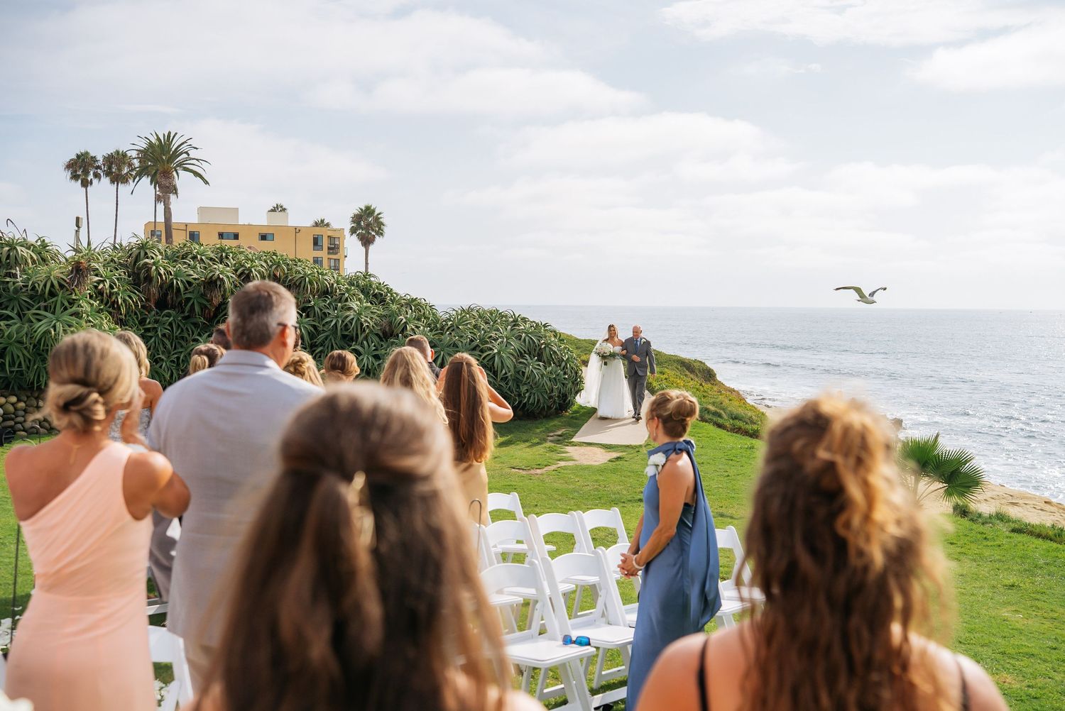 Bride walks down the aisle with escort at an outdoor oceanfront wedding ceremony as guests watch from white chairs.