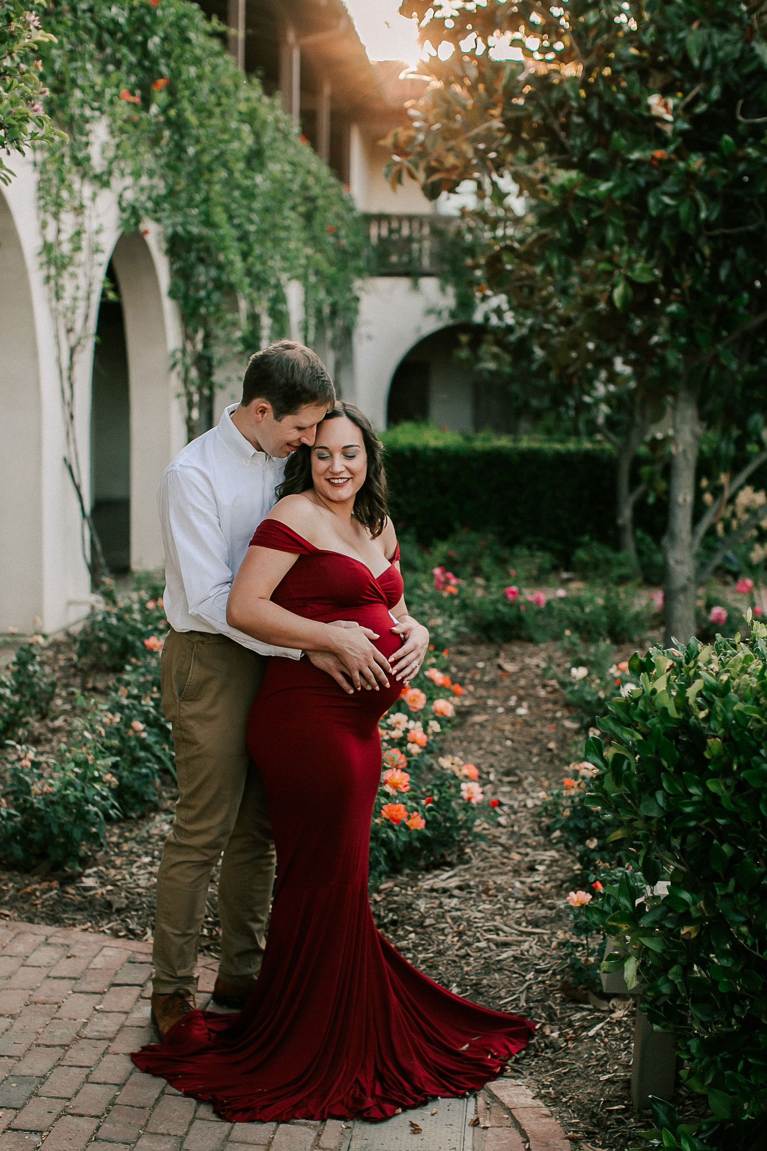 Pregnant woman in red off-shoulder gown with partner in garden courtyard during golden hour maternity photo session.