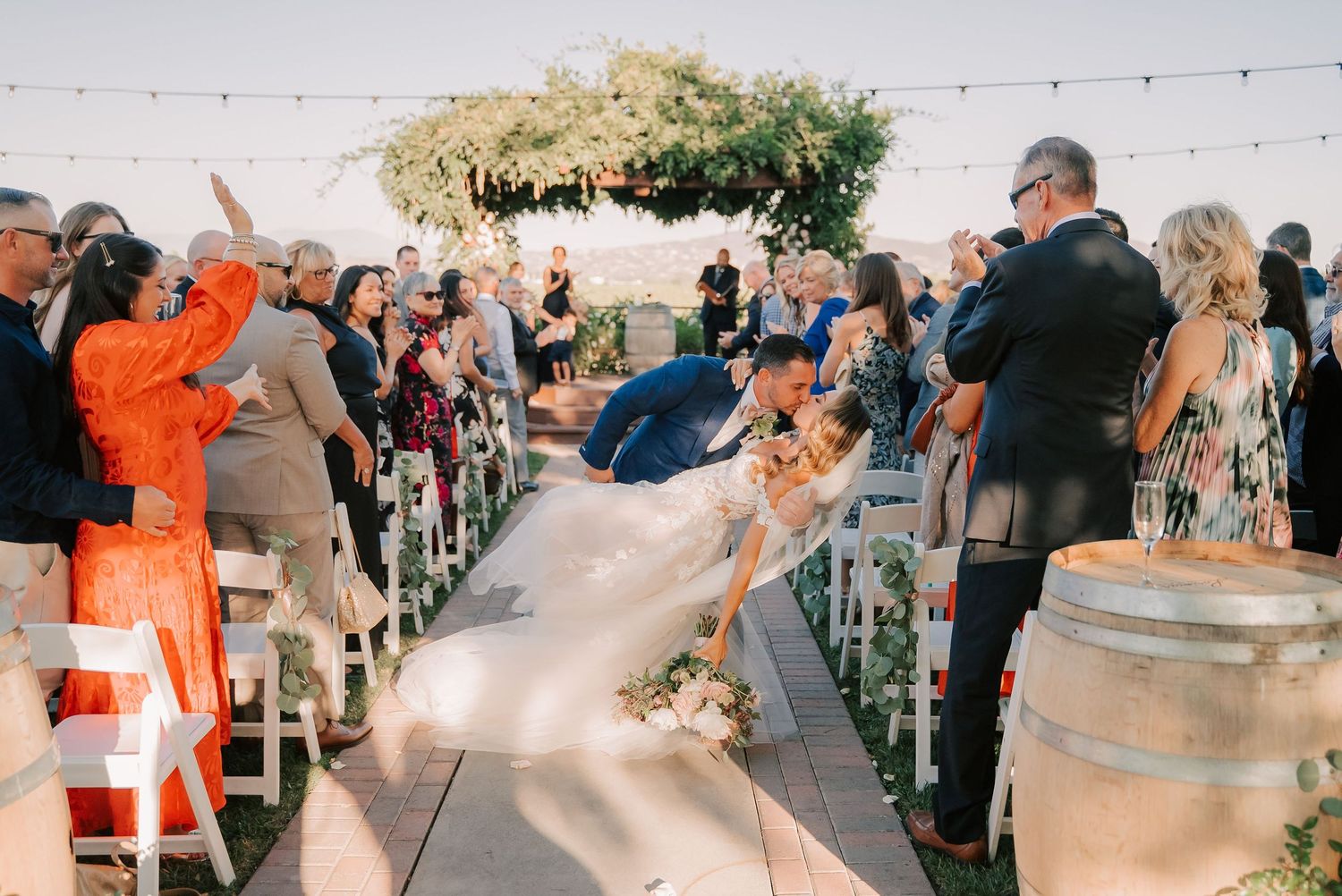 Couple shares romantic dip kiss at outdoor wedding ceremony aisle surrounded by guests and floral arch.