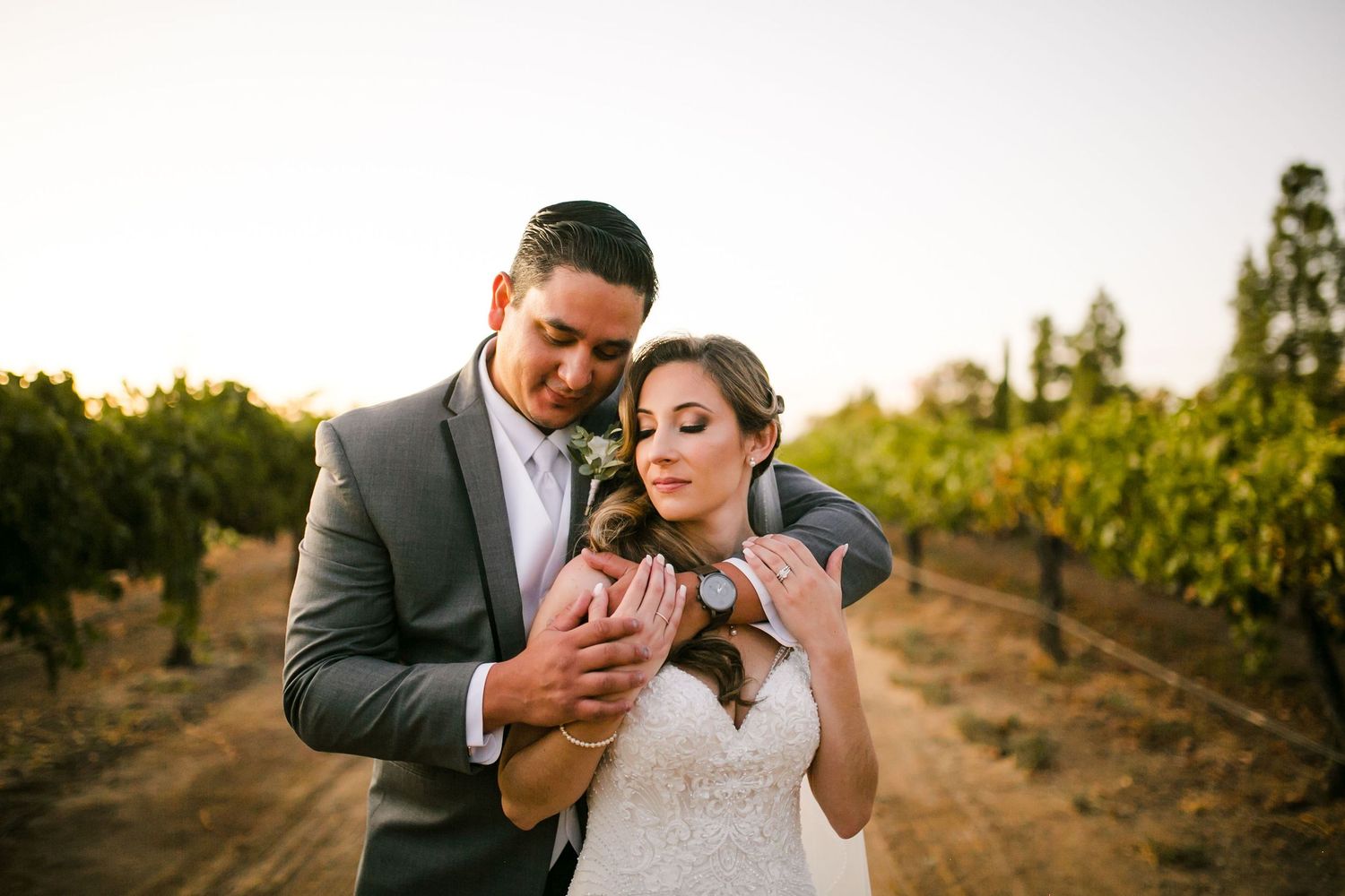 Bride and groom holding a small puppy in a vineyard at golden hour, wearing wedding attire.