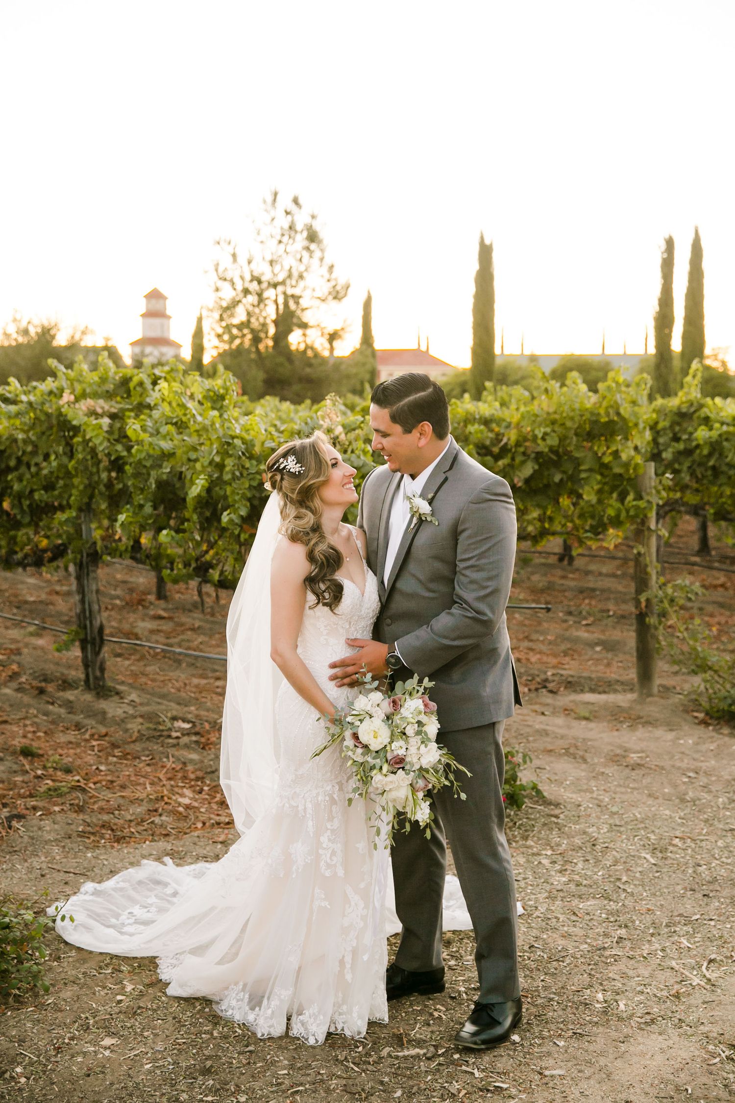 Bride and groom share a romantic moment in a sunlit vineyard, bride in lace gown with bouquet, groom in gray suit.