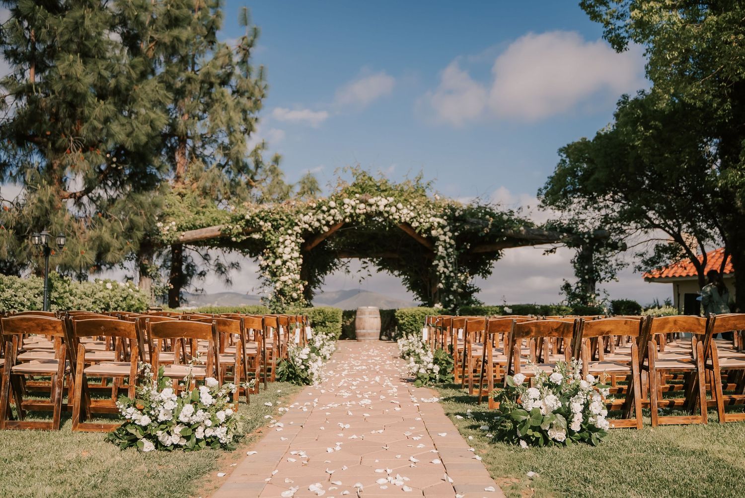 Elegant outdoor wedding ceremony setup with wooden chairs, floral arch, and petal-lined aisle under blue skies.