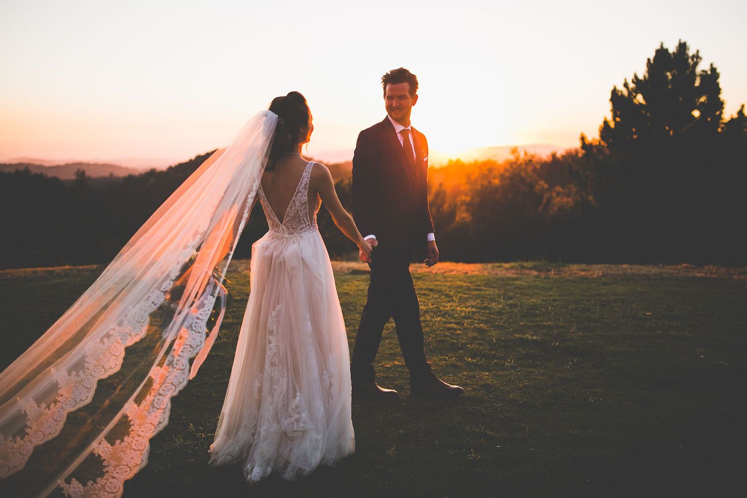 Bride and groom holding hands at sunset on a green lawn, bride wearing lace wedding gown with long veil.