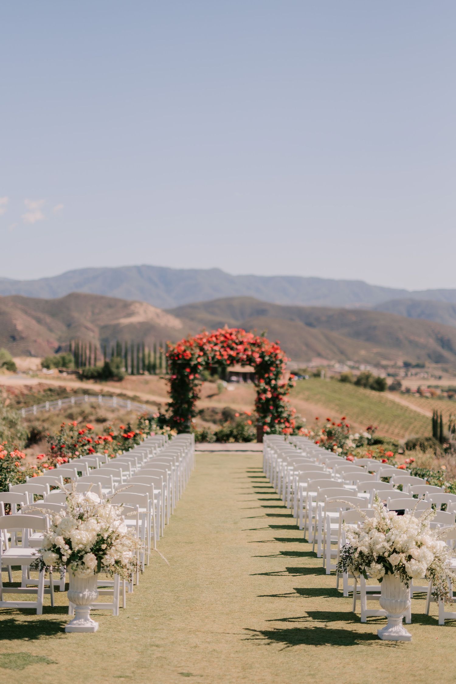 Outdoor wedding ceremony setup with white chairs, floral arch, and mountain vineyard backdrop under clear blue sky.