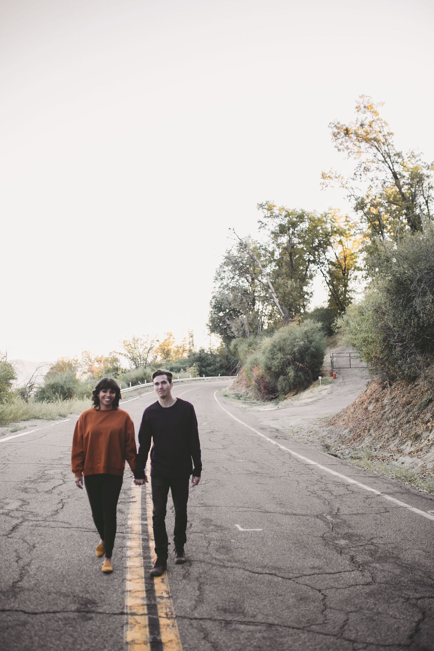 Couple holding hands walking on a winding road surrounded by autumn trees at dusk.