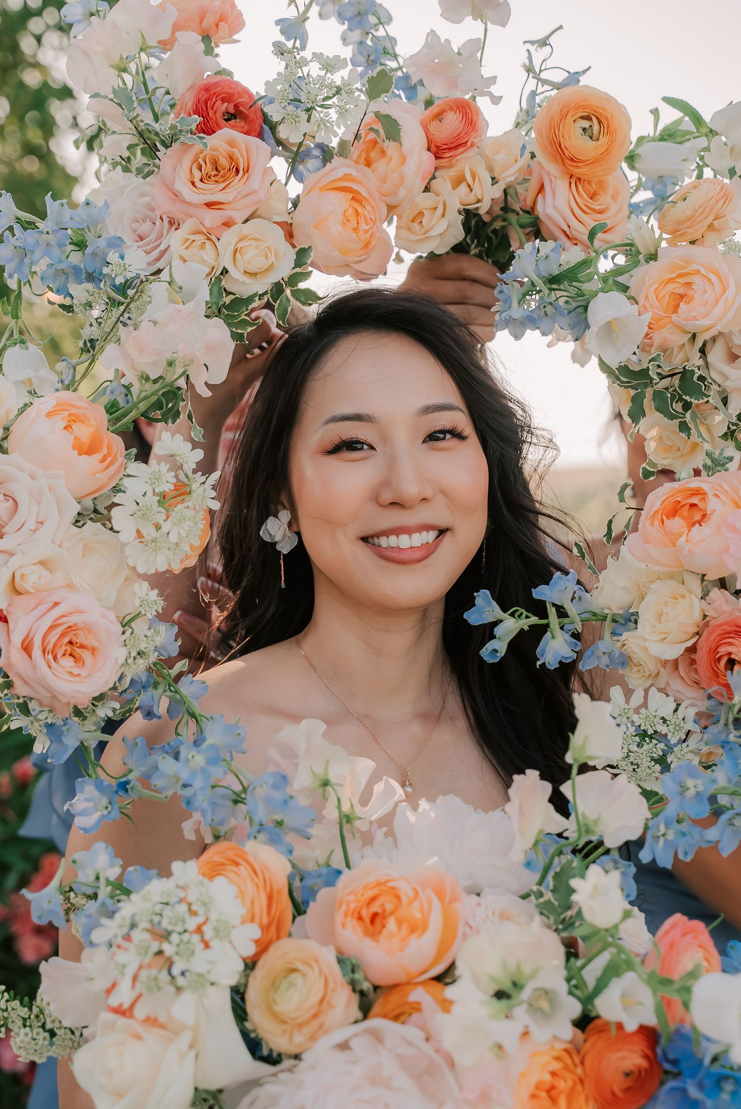 Smiling bride framed by a circular floral arrangement of peach roses, blue hydrangeas, and white blooms.