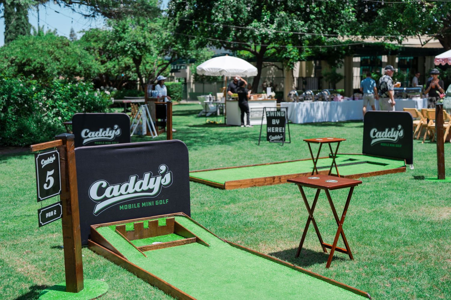 Caddy's mini golf course setup on green turf at an outdoor event with wooden signs and tables.
