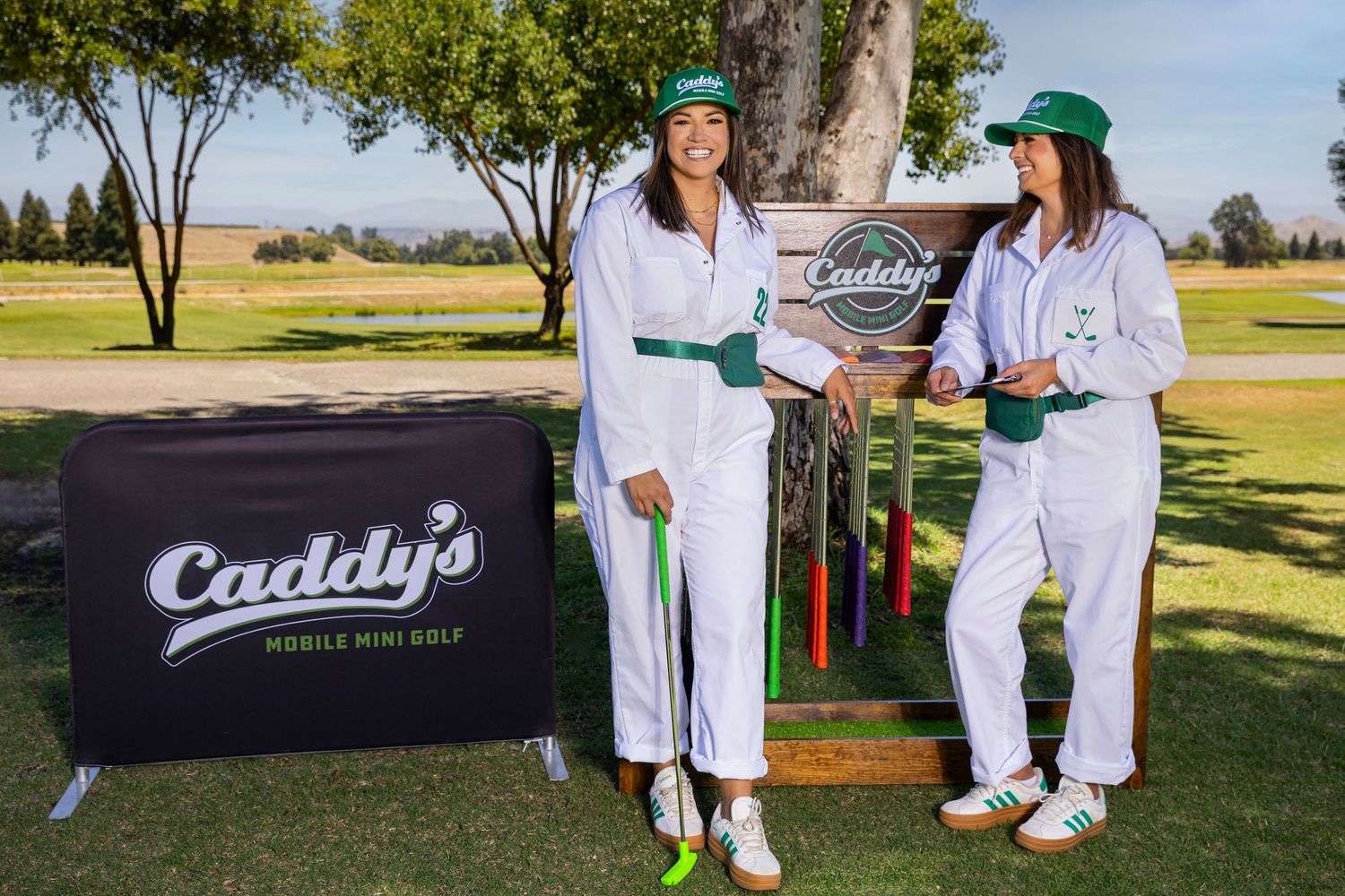 Two women in white uniforms pose with golf clubs at Caddy's Mobile Mini Golf outdoor setup on a sunny day.