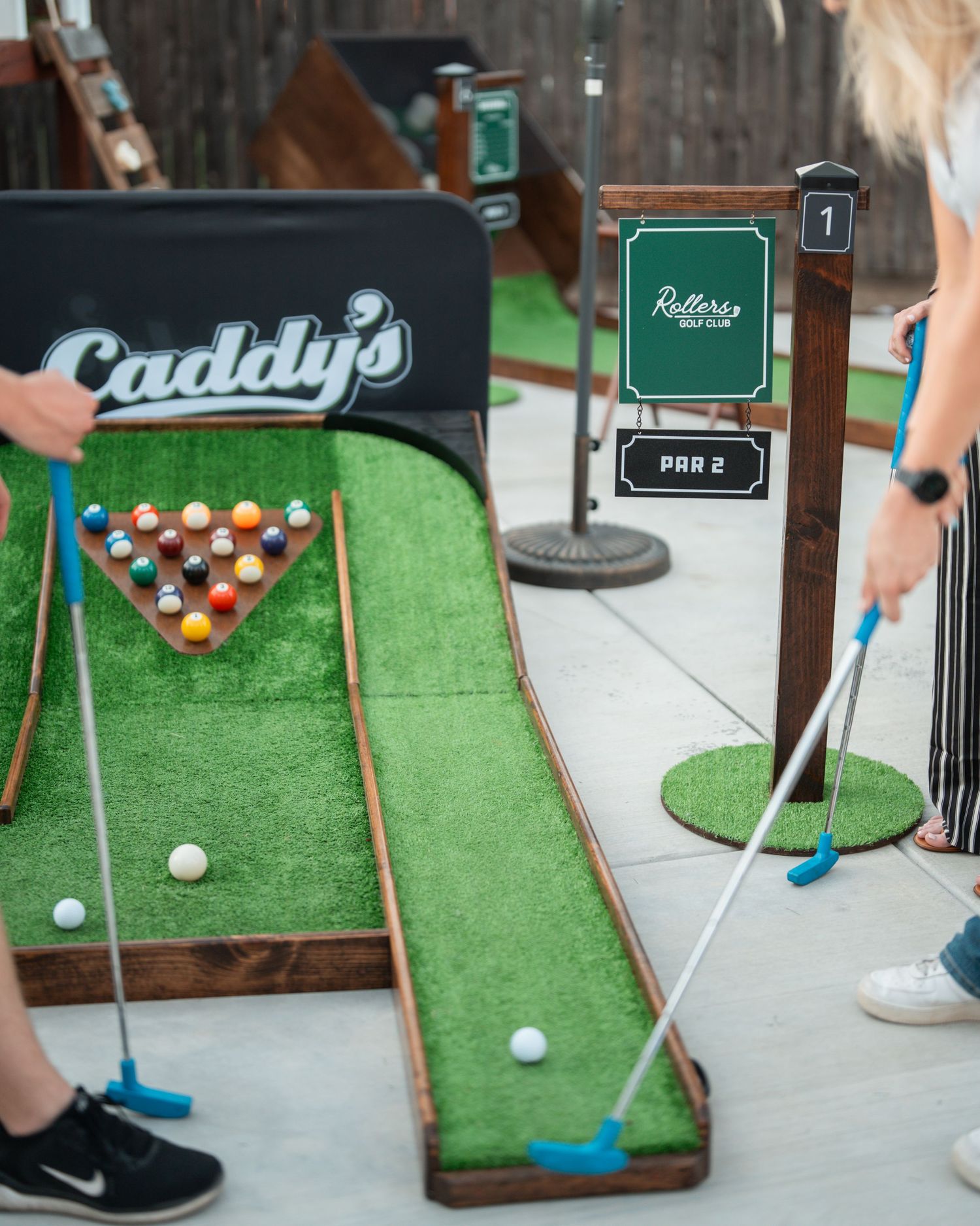 Two players putting at Caddy's Mobile Mini Golf station 1, par 2 hole indoors.