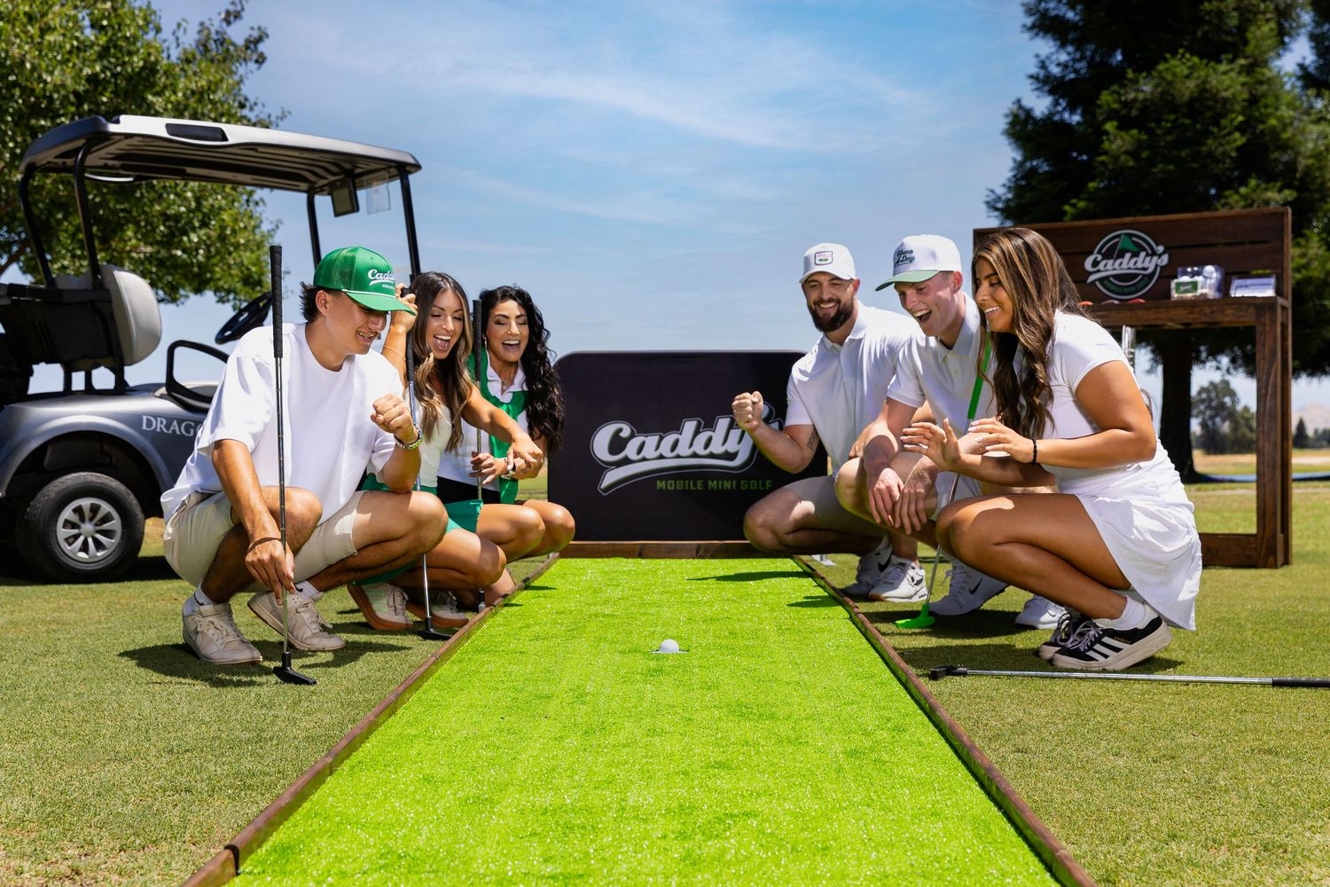Group of six friends crouch around a golf ball on a Caddy's branded putting green outdoors.
