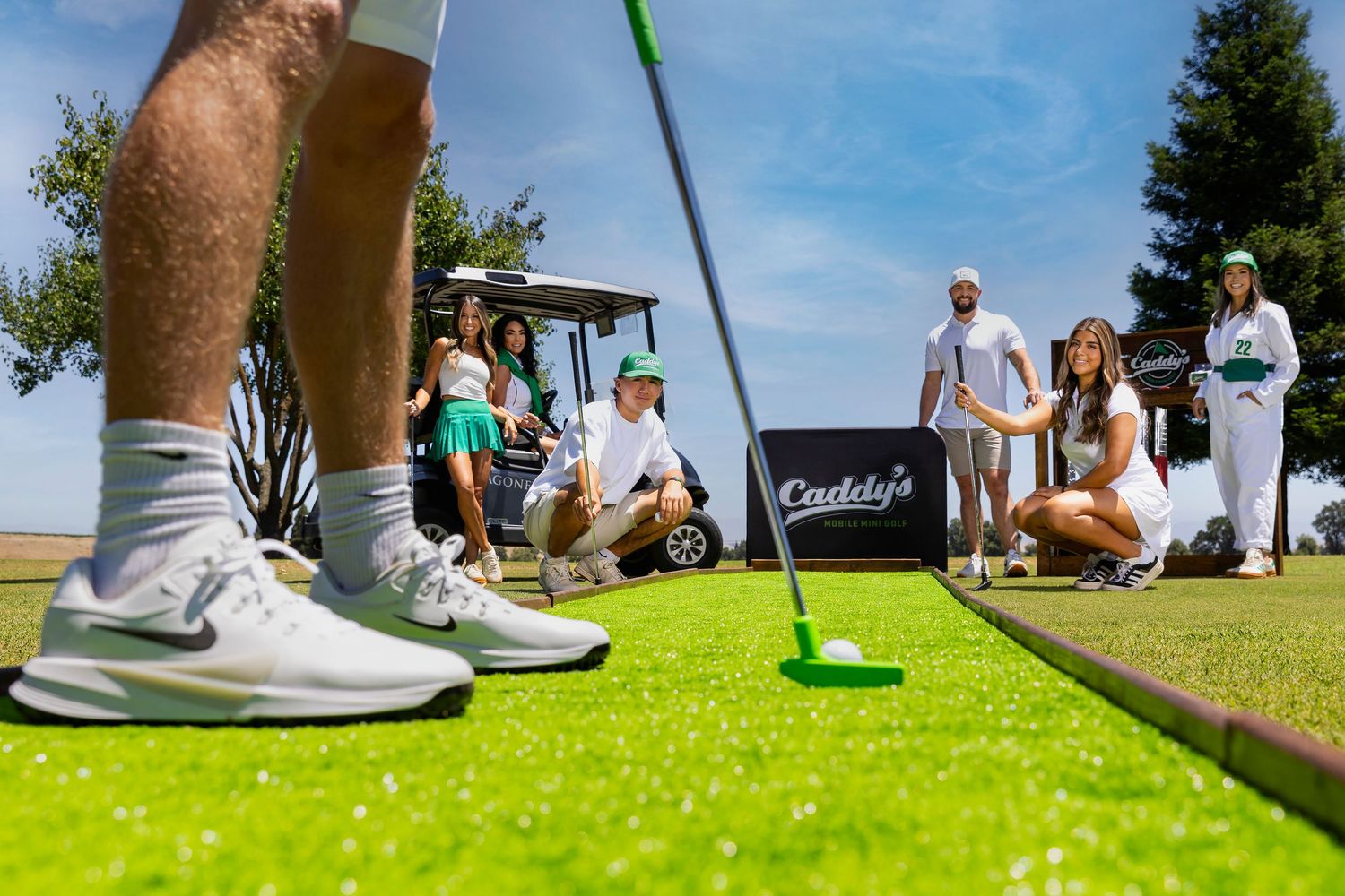 Golfer putts on green at Caddy's golf event with spectators and golf cart in background on sunny day.
