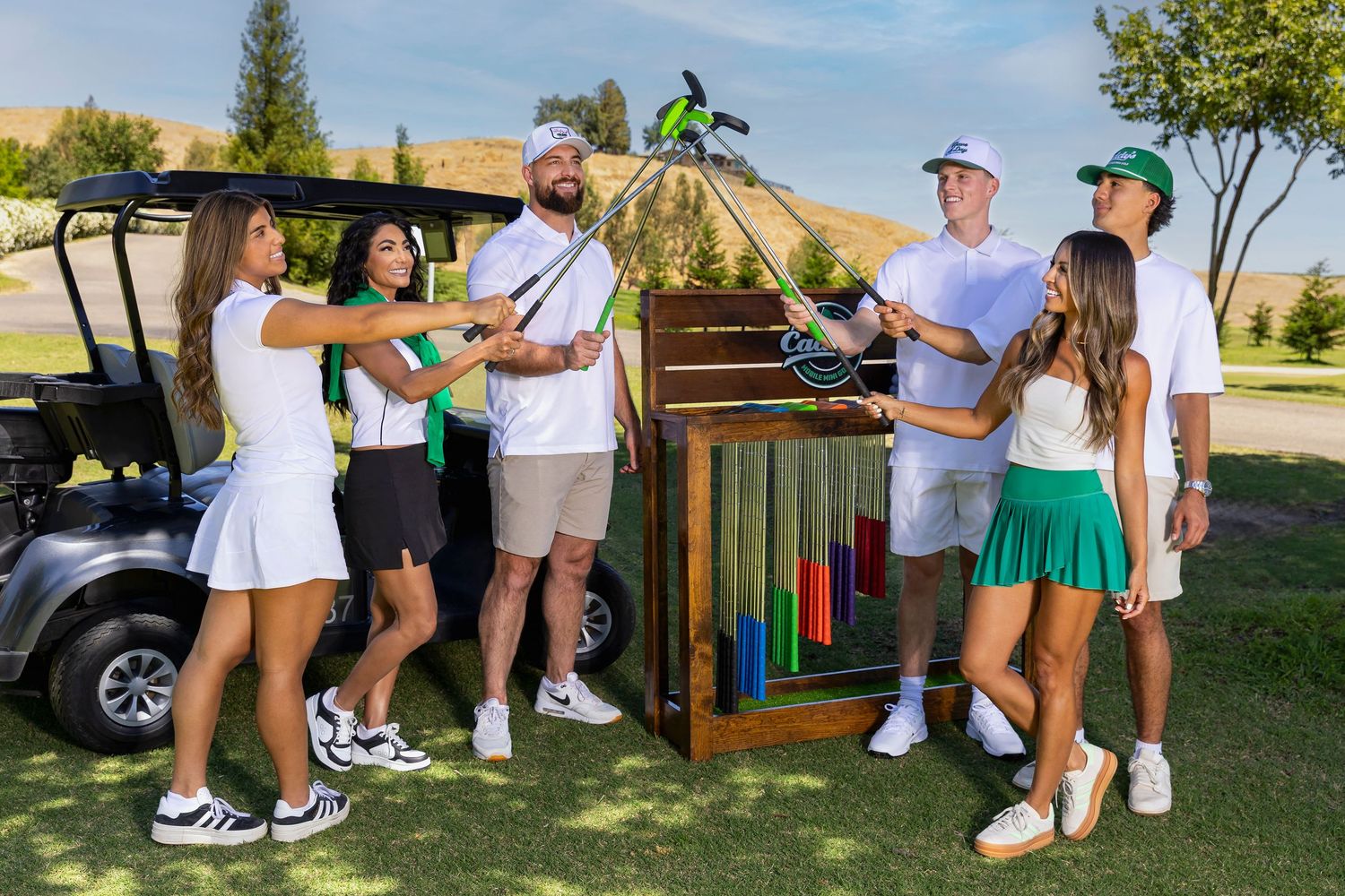 Group of friends gathered around a colorful Caddy's mini golf obstacle near a golf cart outdoors.