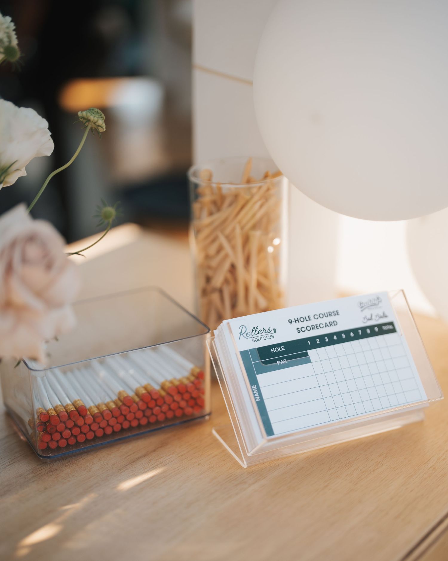Mini golf scorecards and colorful matchsticks in acrylic holders displayed on a white table with flowers.