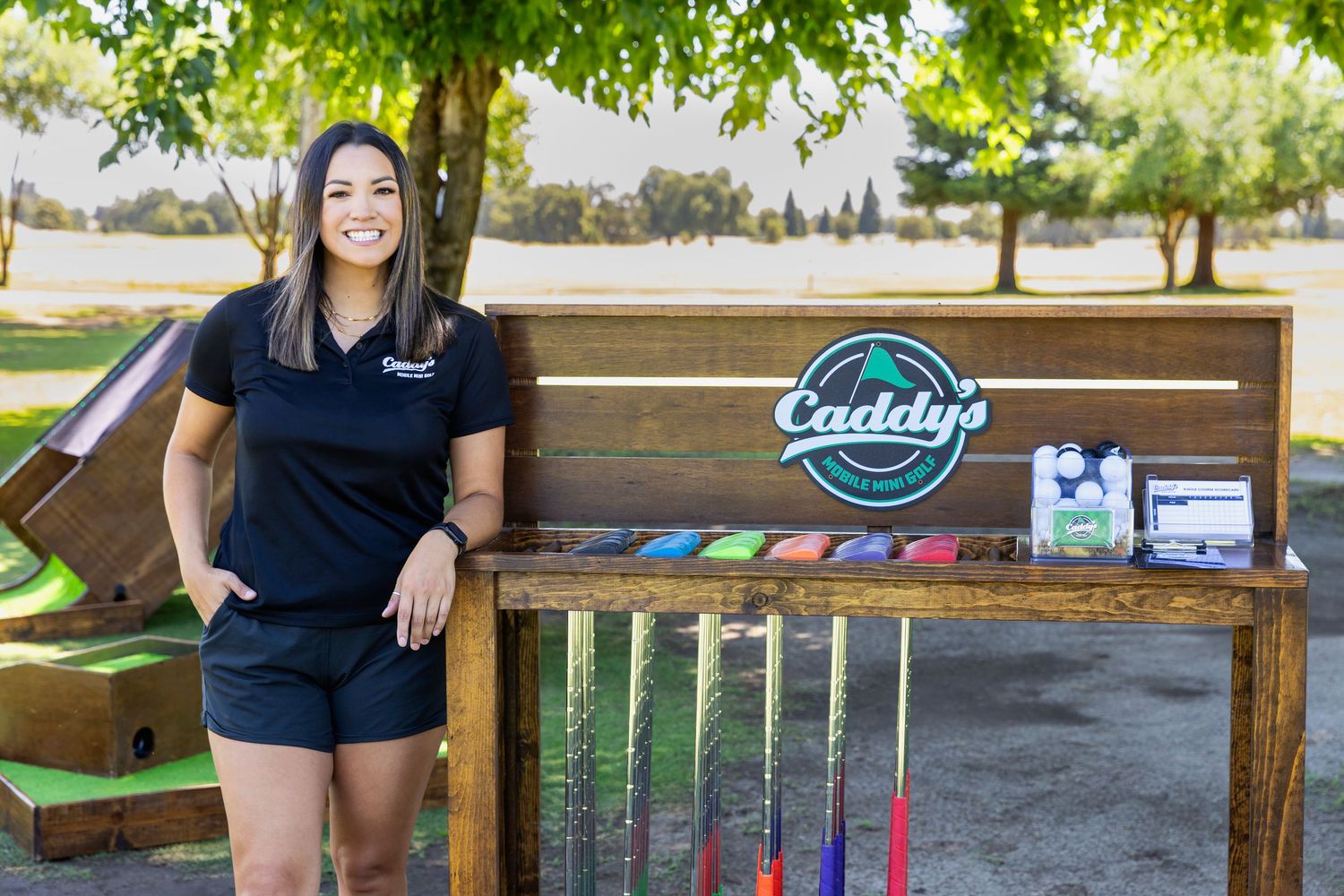 Smiling woman in black Caddy's uniform leaning against a wooden club rack at an outdoor golf event.