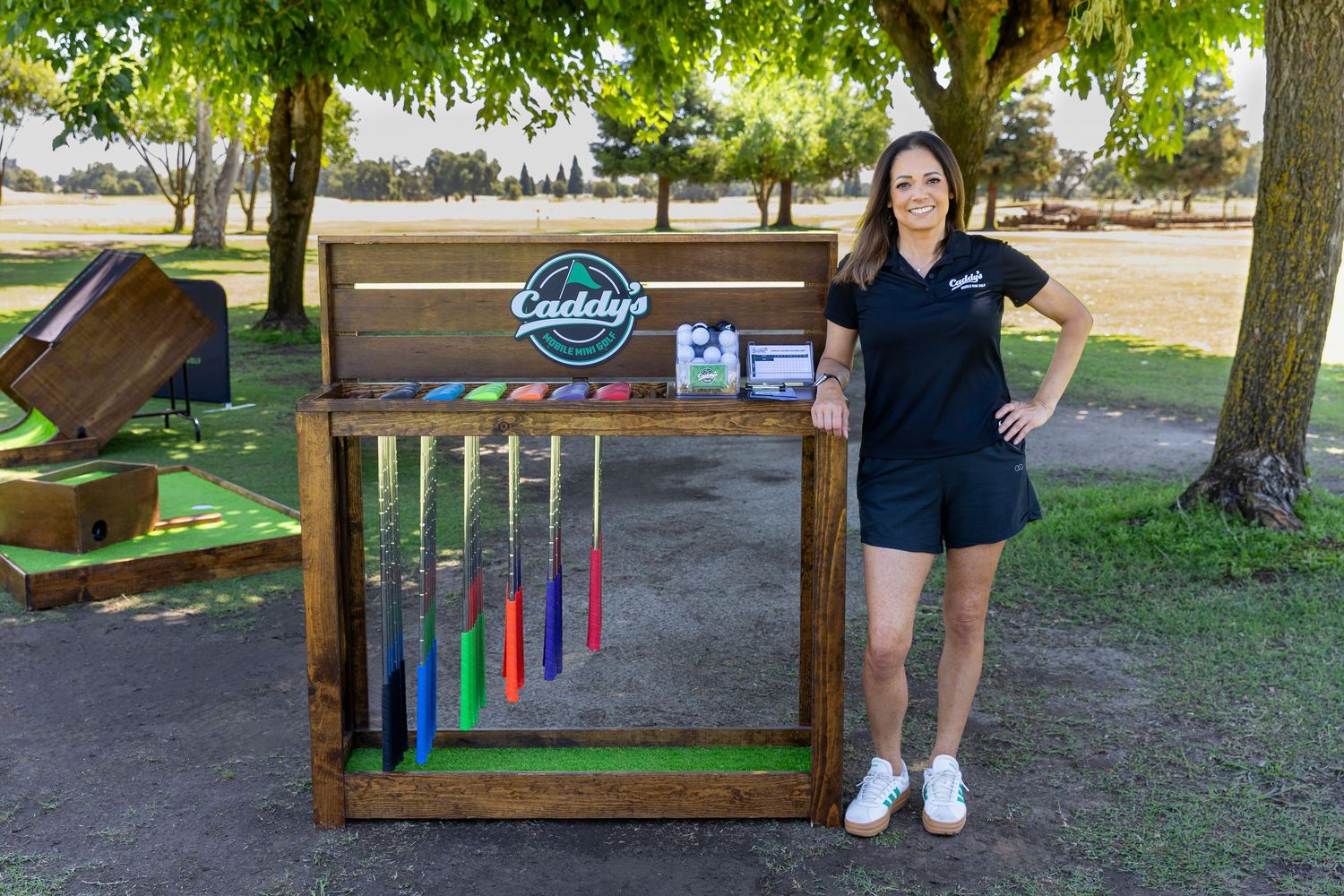 Woman in black Caddy's polo shirt posing beside a wooden mini golf club rack outdoors.