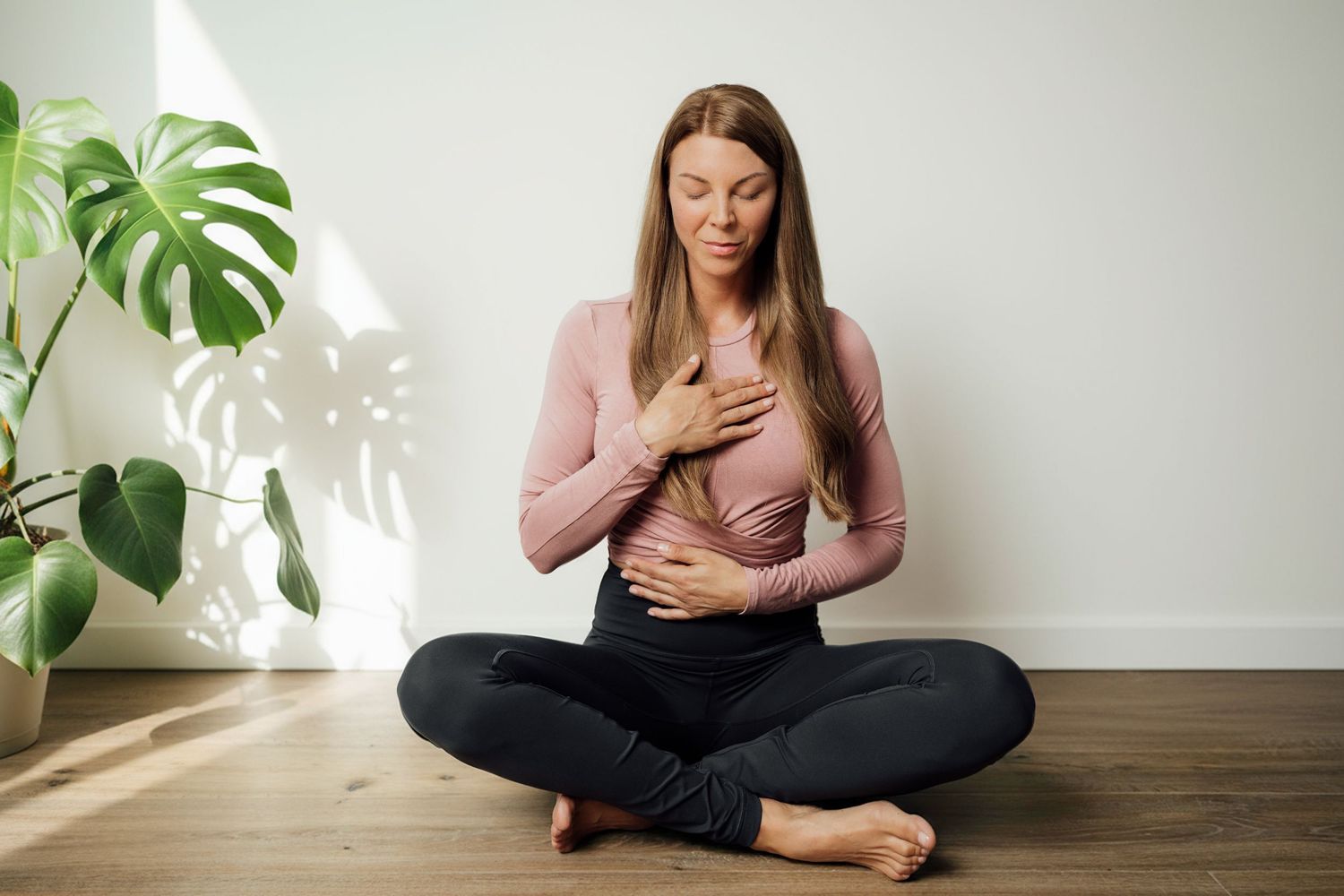Person sitting cross-legged on wooden floor with large monstera plant in background.