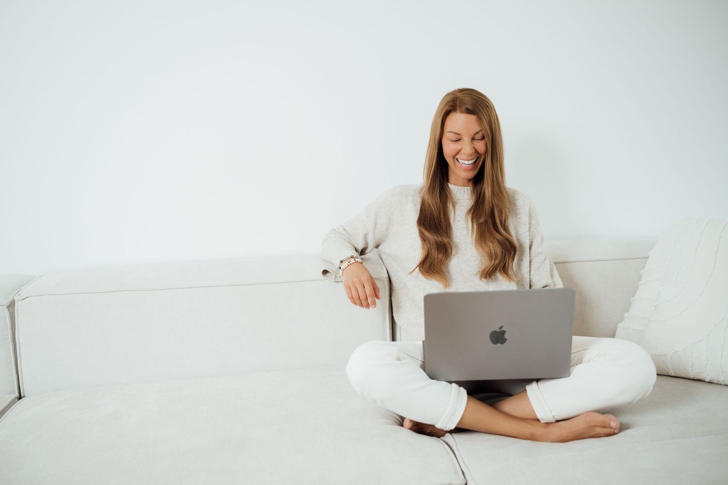 Person working on laptop while relaxing on white couch in minimalist room setting.