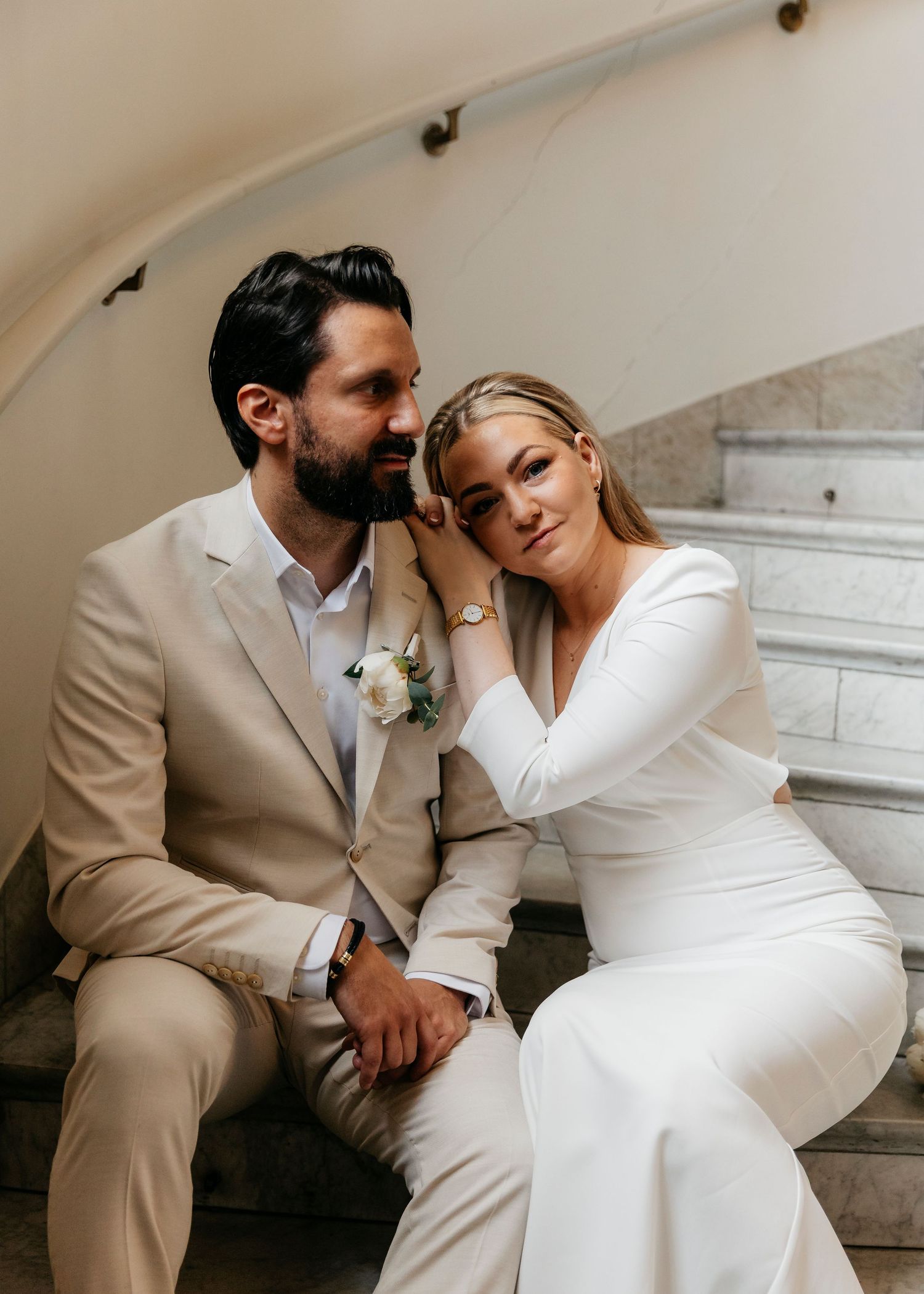A couple in elegant white attire shares romantic poses on marble stairs.