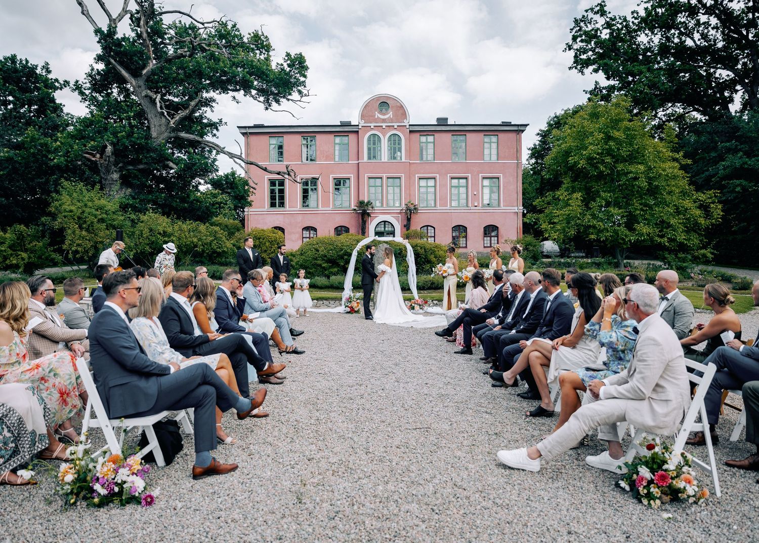 Wedding ceremony seating arrangement on gravel with pink building in background.