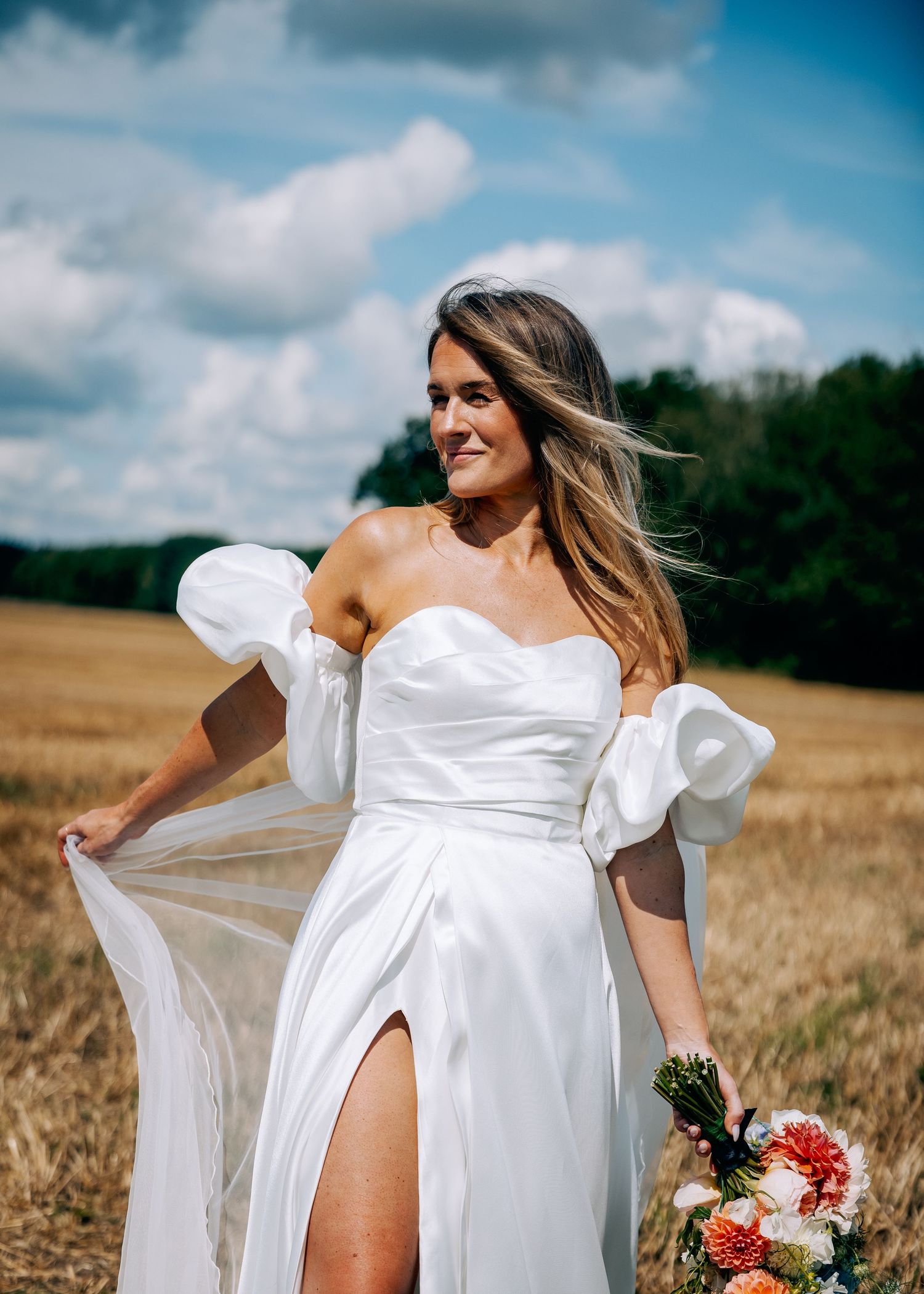 Bride in flowing white dress holding wildflower bouquet in wheat field.