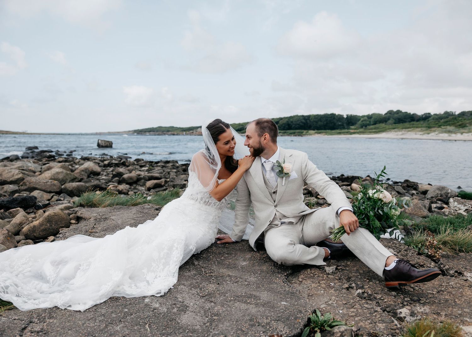 Wedding couple poses playfully along rocky coastline with water views.