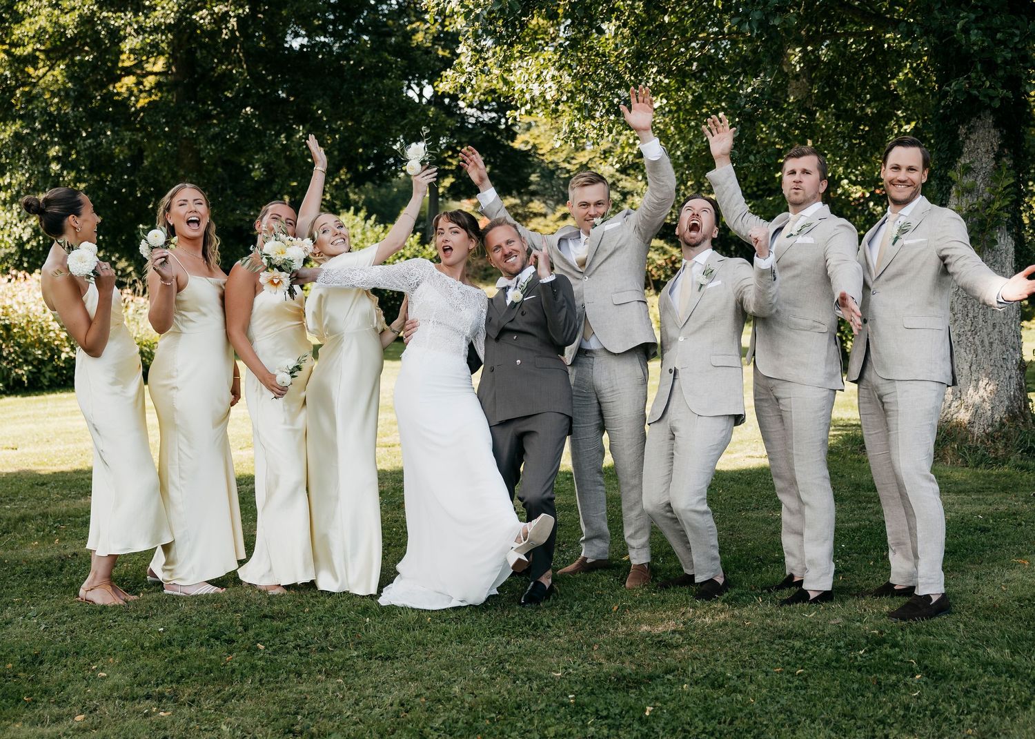 Wedding party in white dresses and gray suits poses playfully on grass.