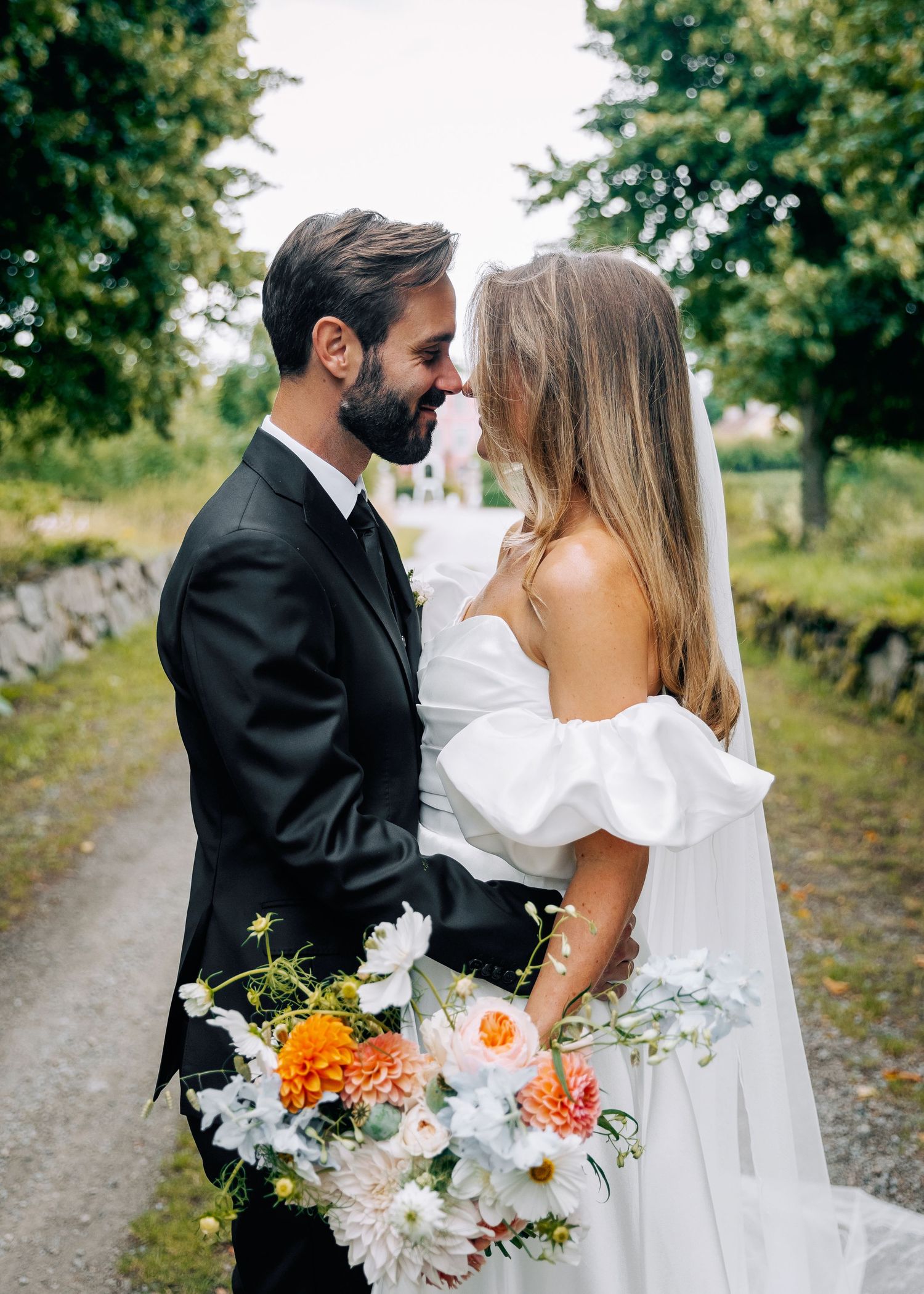 Romantic wedding couple embraces on a tree-lined path while holding a colorful summer bouquet.
