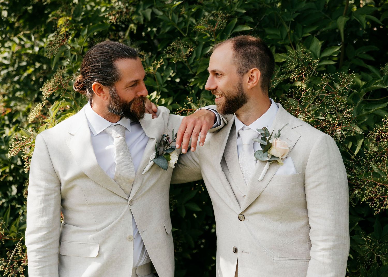 Groomsmen in cream suits sharing candid moments against green foliage.