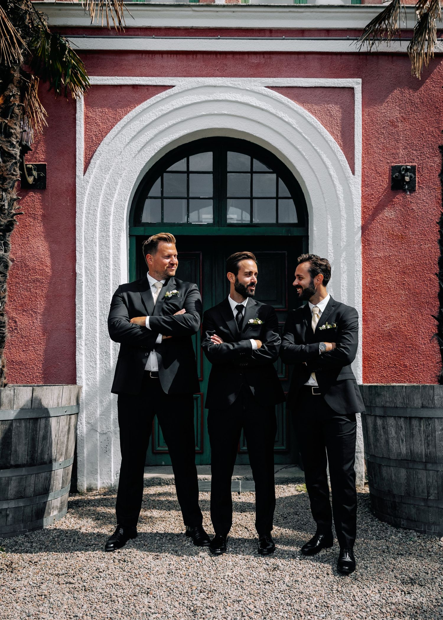 Three groomsmen laughing together under arched doorway of pink building.